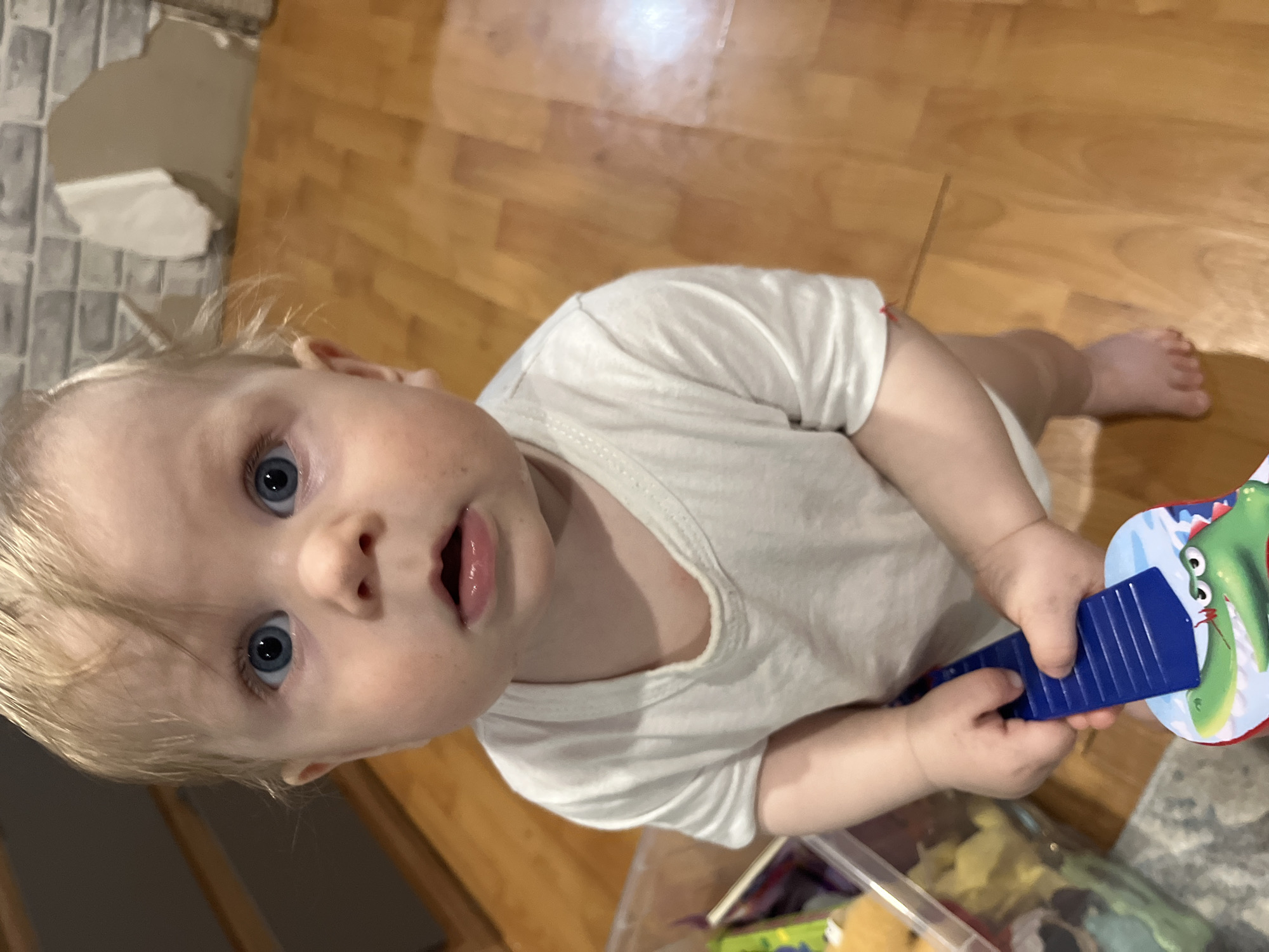 A young child with blonde hair and blue eyes is looking up at the camera. The child is wearing a white t-shirt and holding a blue and multi-colored toy in their hands. The child appears to be indoors on a wooden floor. In the background, there's a partially damaged wall. A plastic container is visible near the child. The location cannot be determined from the image.