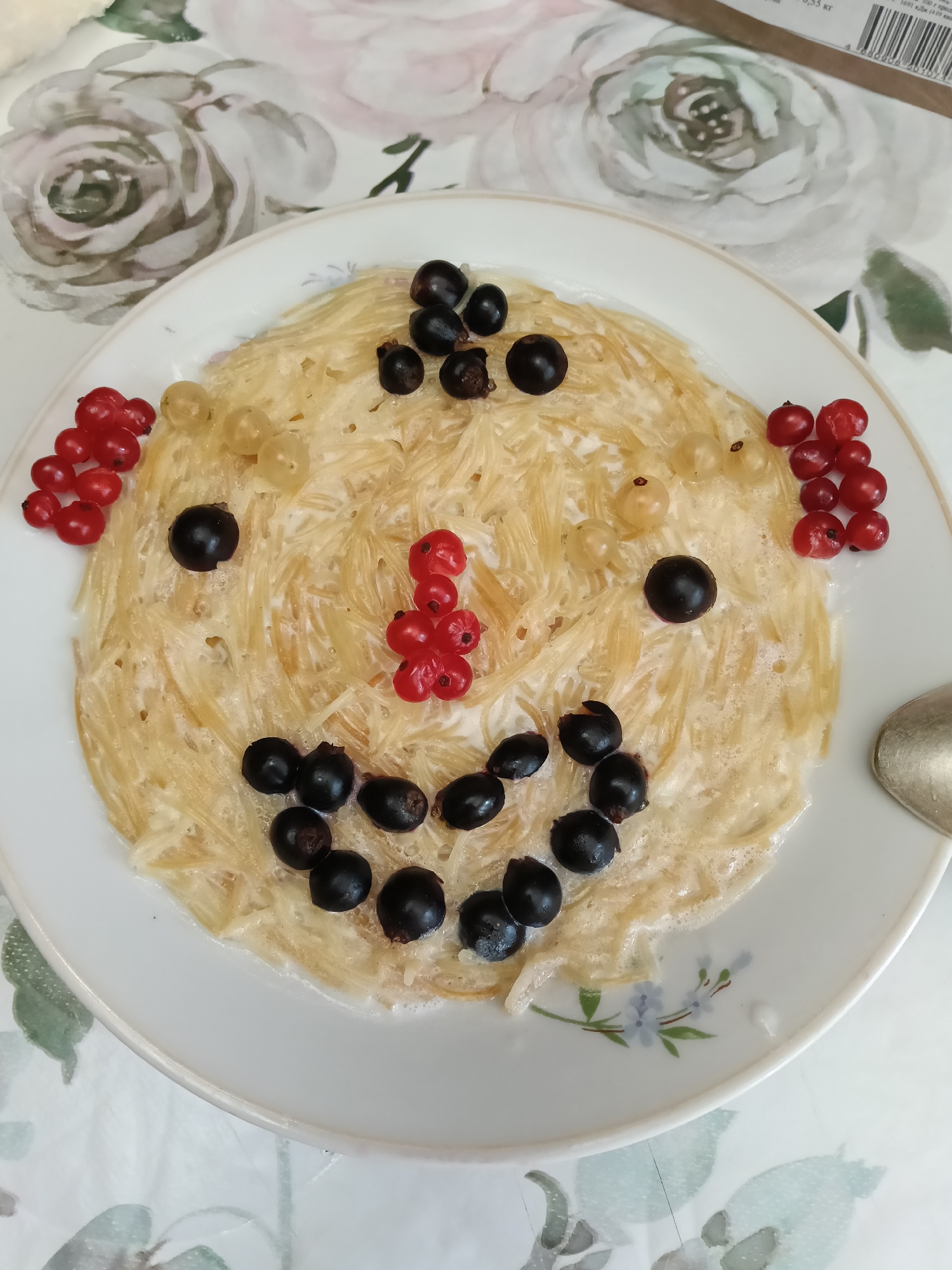 A close-up, overhead shot shows a plate of food resembling a smiley face, located in the city of Krasny Mayak, Russia. The main dish consists of pale yellow noodles coated in a creamy white sauce, arranged in a circular mound on a white plate. The "face" is decorated with black currants for eyes and a mouth, white currants for cheeks, and red currants forming a nose and the sides of the smile. The plate has a subtle floral pattern near the bottom edge. The background is a table covered with a floral-patterned tablecloth, featuring light-colored roses. A silver spoon is visible to the right of the plate.