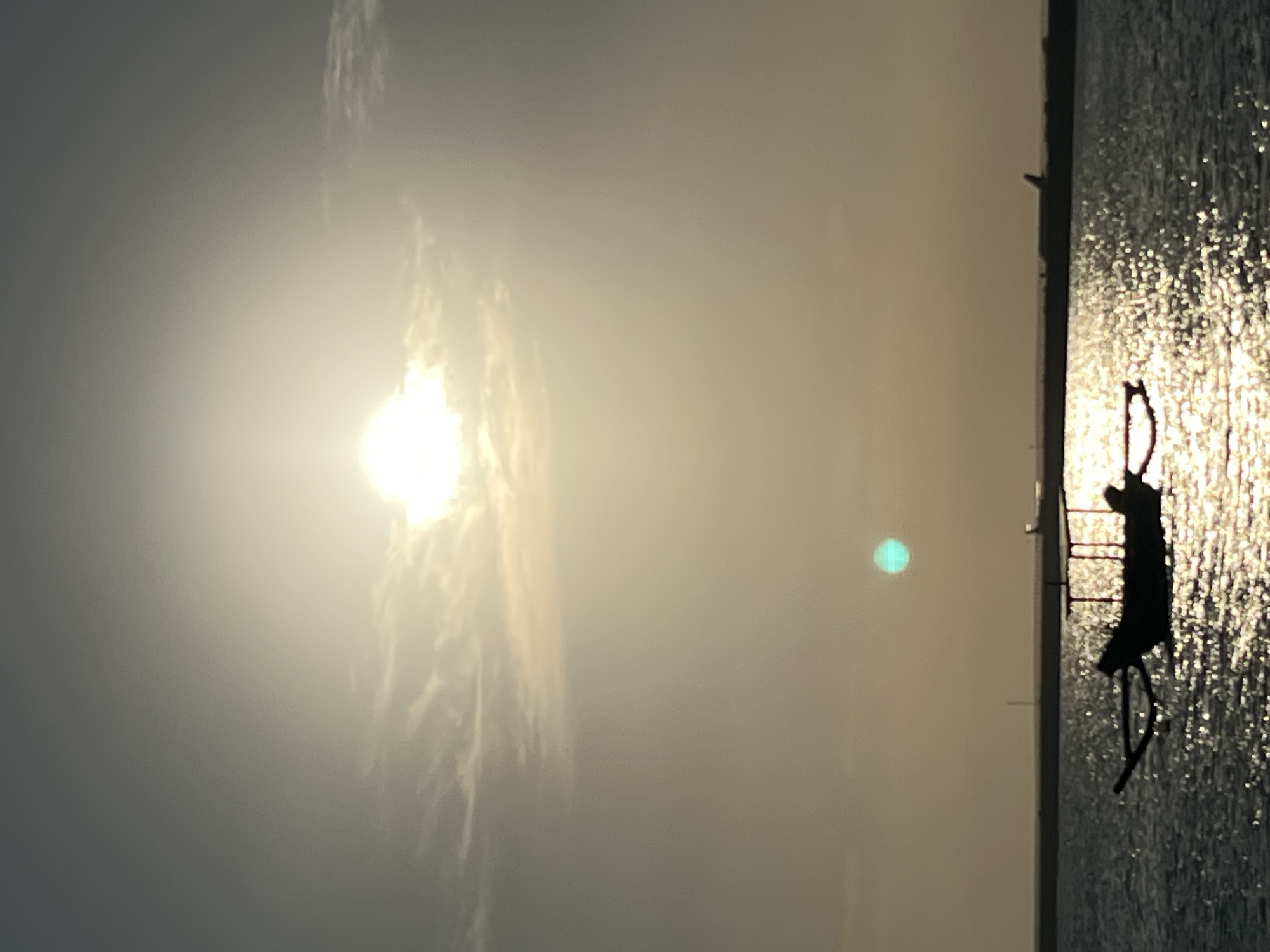 The image depicts a serene scene at Kuta beach, Indonesia, during a sunset. The sun, a bright orb with a halo of light, is partially obscured by faint clouds in the upper portion of the frame. Below the sun, a turquoise orb of light hangs in the hazy sky. The foreground is dominated by the ocean, with its surface shimmering with the reflection of the setting sun. A traditional Indonesian fishing boat, silhouetted against the bright water, is visible in the mid-ground. The boat appears to be anchored or stationary. In the background, a distant landmass stretches horizontally, possibly a coastline with some structures or a breakwater. Two faint airplane silhouettes can be discerned in the distance, suggesting the proximity of an airport. The overall atmosphere is tranquil and idyllic, characteristic of a tropical sunset.