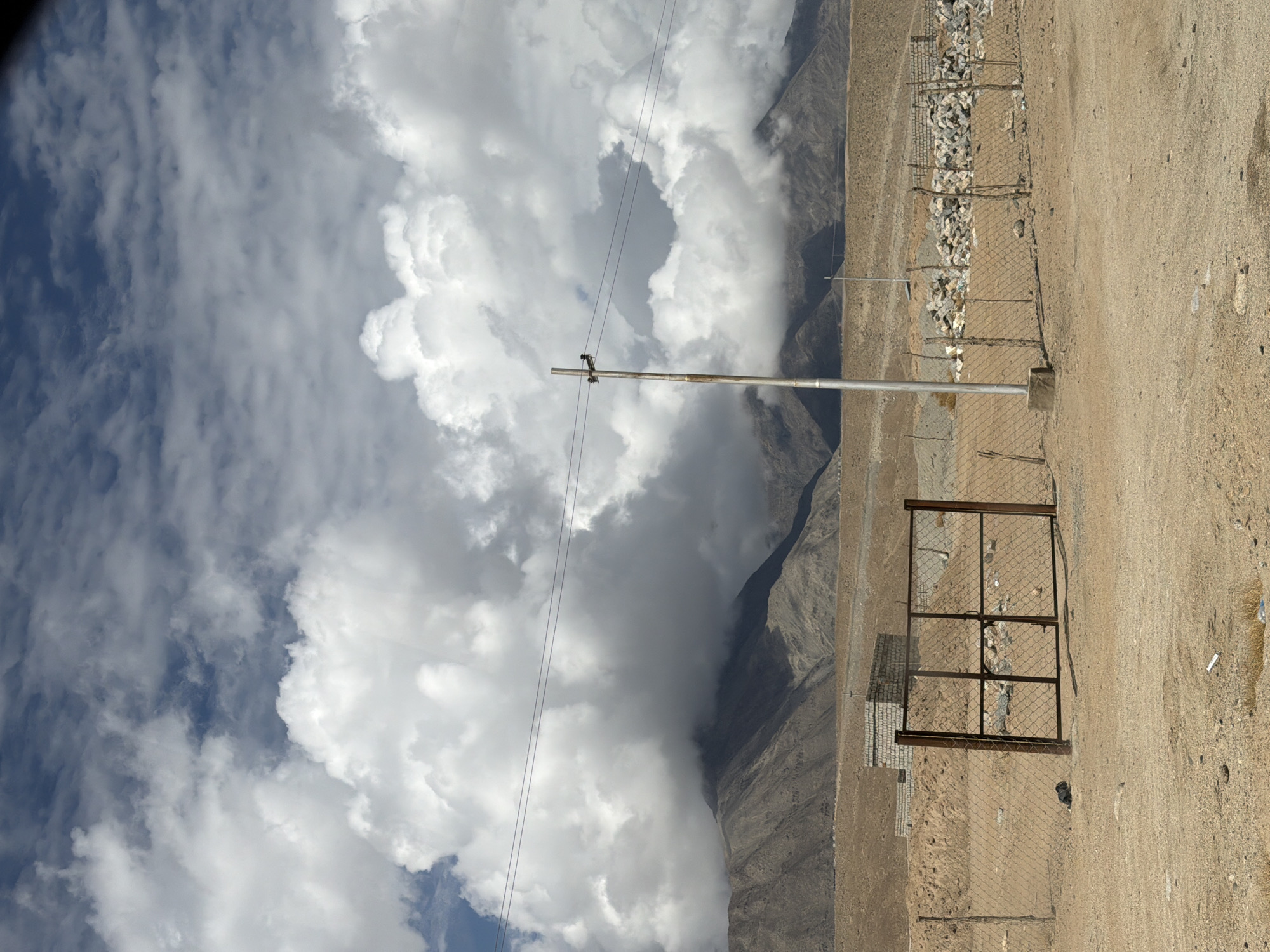The image depicts a wide, arid landscape under a cloudy sky. In the foreground, a sandy ground is visible with scattered rocks and debris. A chain-link fence with a metal gate, appearing rusted, stretches across the middle ground. Behind the fence, a small, unfinished brick structure is situated, accompanied by piles of rocks. The background is dominated by rugged, brown mountains partially obscured by large, white clouds. A utility pole with two wires is positioned to the right of the frame, and another shorter pole is visible further back. The scene appears to be located in the city of Chumkar, India, given the arid environment and mountainous terrain characteristic of the region.