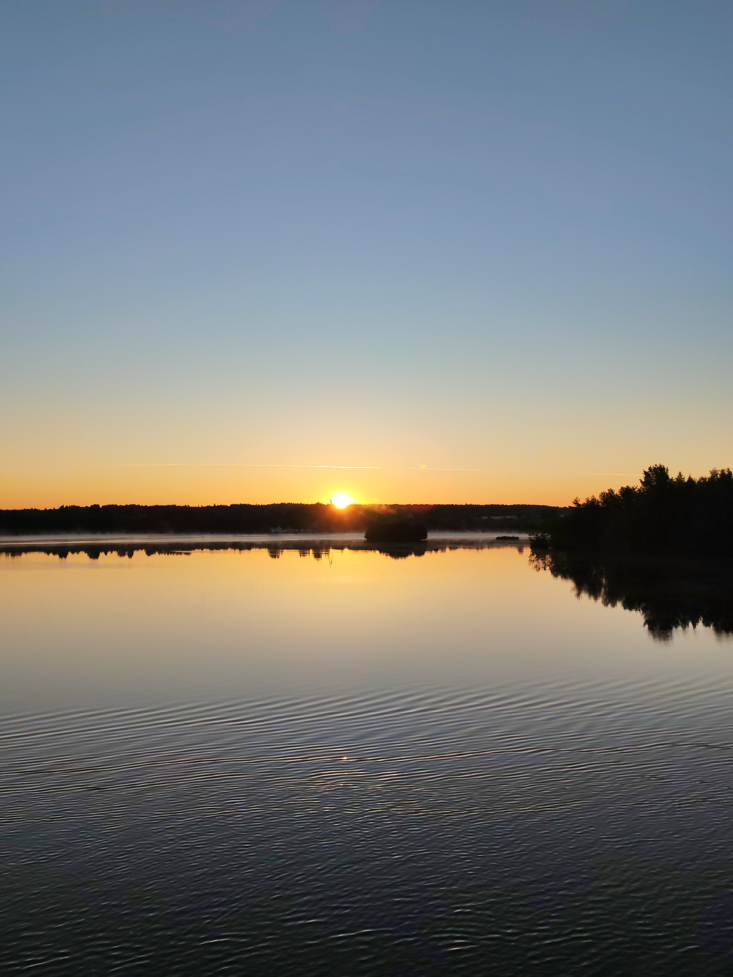 The image depicts a serene lake at sunrise. The sun is just cresting the horizon, casting a warm orange glow across the sky and reflecting on the calm water. A dense line of trees forms a dark silhouette along the distant shoreline. In the foreground, the water has gentle ripples, creating a textured surface. The sky transitions from a pale blue at the top to a vibrant orange near the horizon. There are no people, animals, or buildings visible in the scene. The location is identified as Devyatiny, Russia.