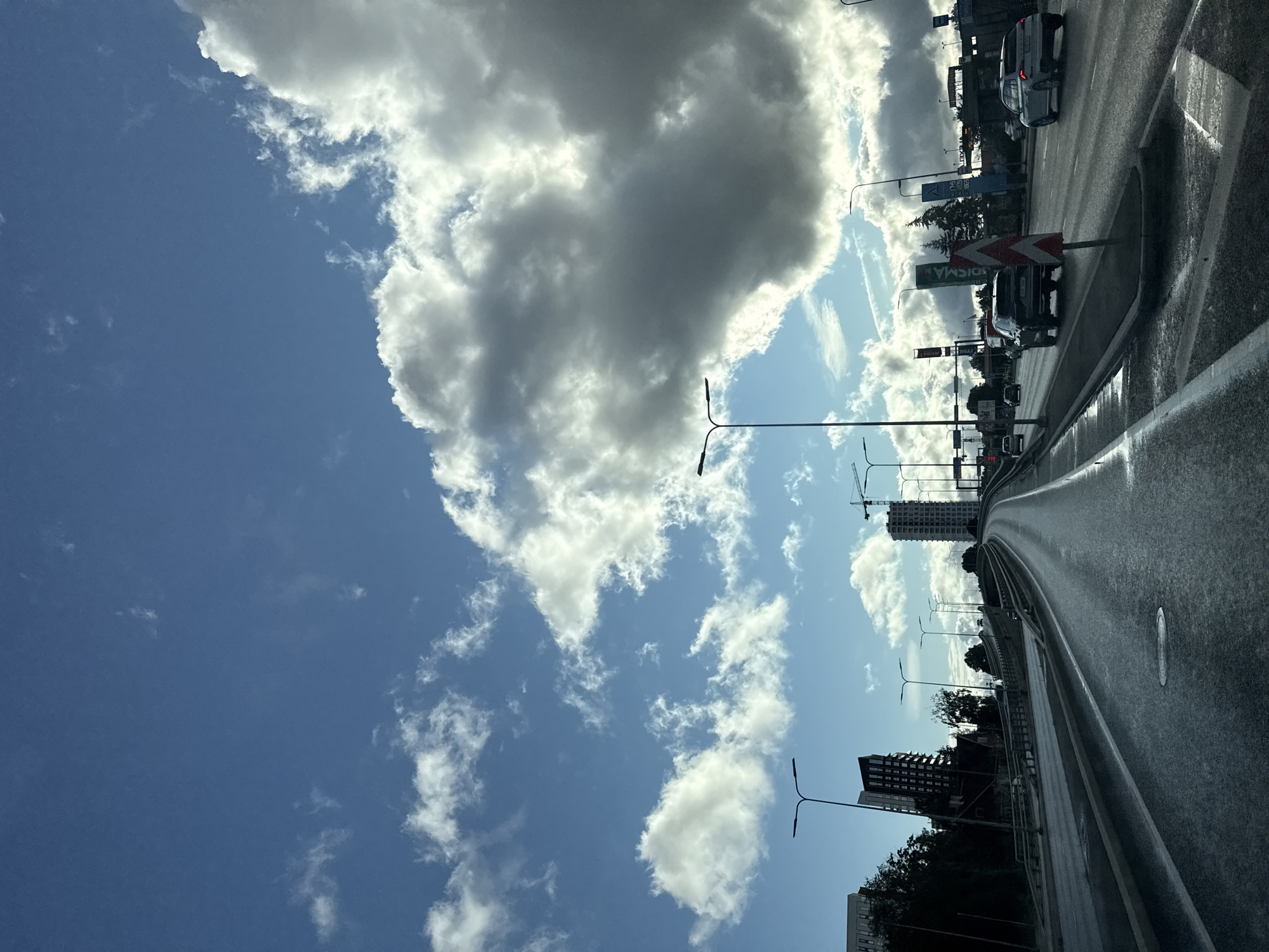 The image depicts a highway in Tallinn, Estonia, under a sky filled with dramatic, cumulus clouds. The highway is wet, suggesting recent rain, and several vehicles are in motion. On the left side of the road, there are modern, dark-colored buildings and trees. On the right side, there are traffic lights, signage, and more buildings in the distance. A construction crane is visible further down the road, indicating ongoing development. The overall impression is a contemporary urban landscape transitioning into a highway, with the weather adding a sense of dynamism to the scene.