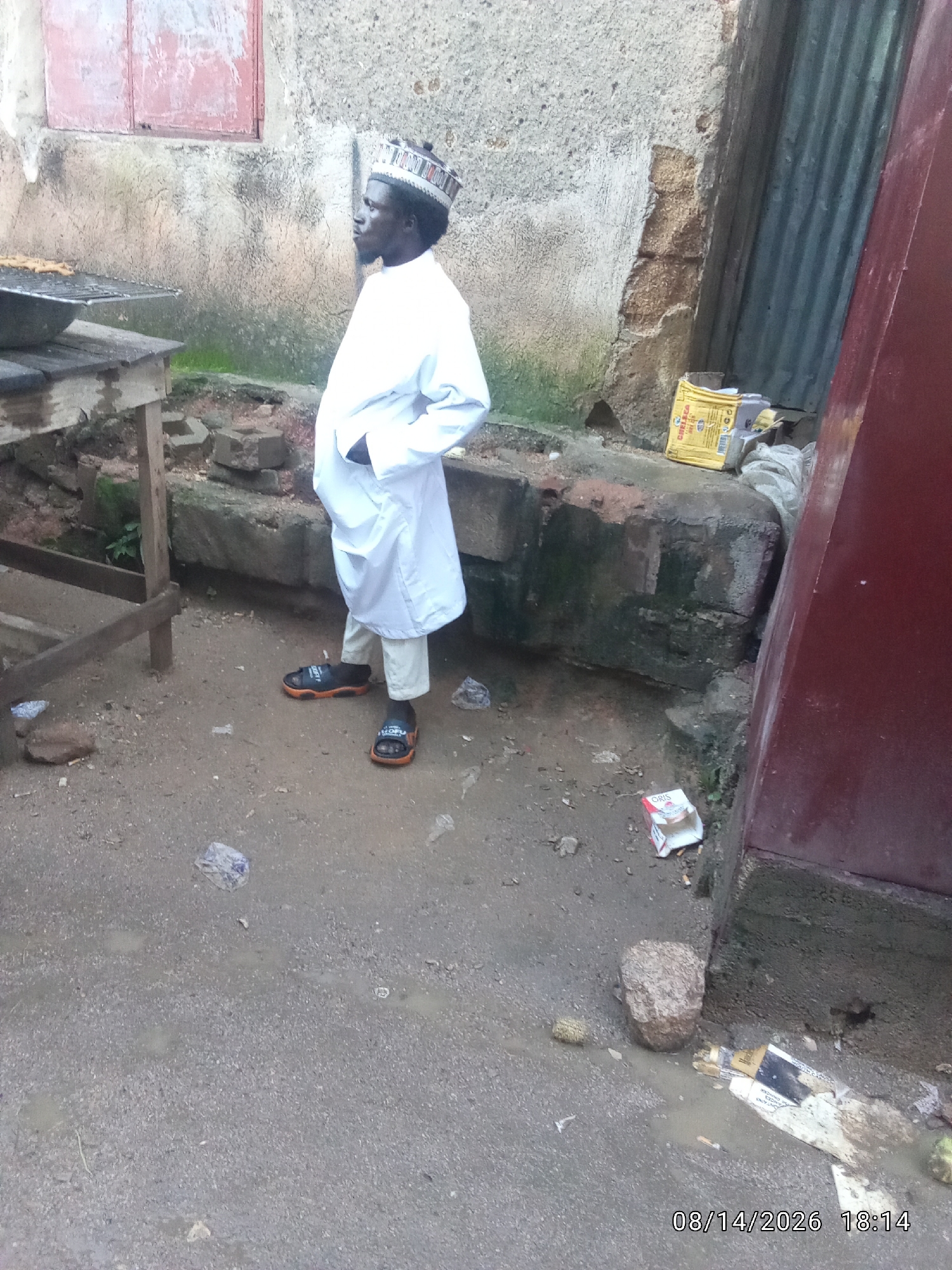 A man in a white robe and a patterned cap stands with his hands in his pockets in front of a dilapidated building. The ground is a mix of dirt and loose gravel, with scattered debris including plastic bags, cigarette butts, and a crushed carton. To the left, a wooden table with a metal rack is visible, suggesting it might be a food stall. In the background, the building's walls are rough and stained, with one section partially covered by corrugated metal sheeting. The scene appears to be an outdoor urban setting in Jos, Nigeria, possibly in a less affluent or unkempt area. A timestamp in the lower right corner reads "08/14/2026 18:14".