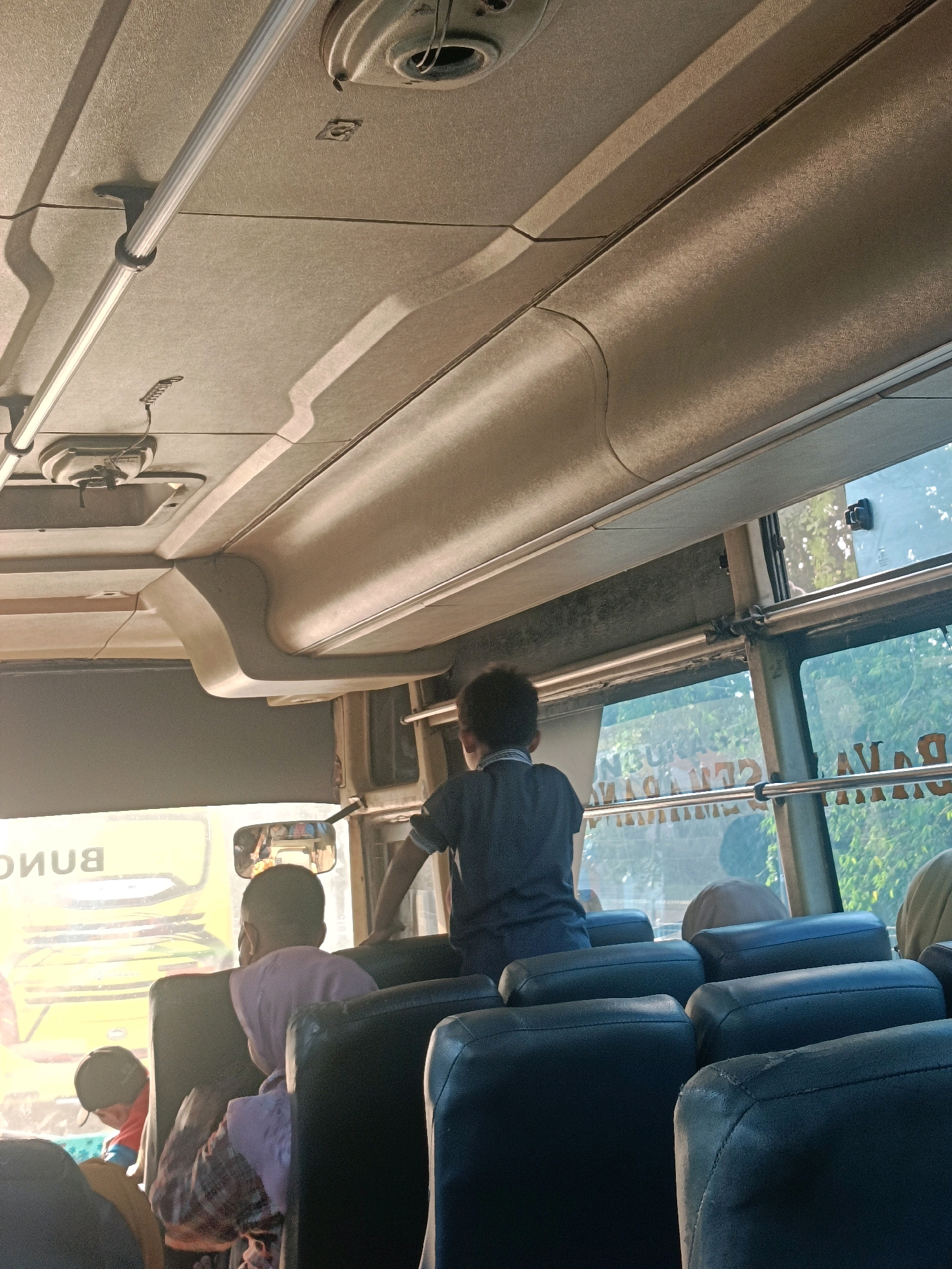 The image shows the interior of a bus, likely in Kedungrejo, Indonesia. The ceiling of the bus is visible, with overhead compartments and light fixtures. Several passengers are seated, facing forward. A young boy in a dark blue shirt stands in the aisle, looking out the window. The windows reveal a blurry view of greenery and possibly other vehicles outside. The seats appear to be dark-colored and upholstered. A mirror reflects some of the passengers in the back. The overall lighting suggests it is daytime, with sunlight entering through the windows.