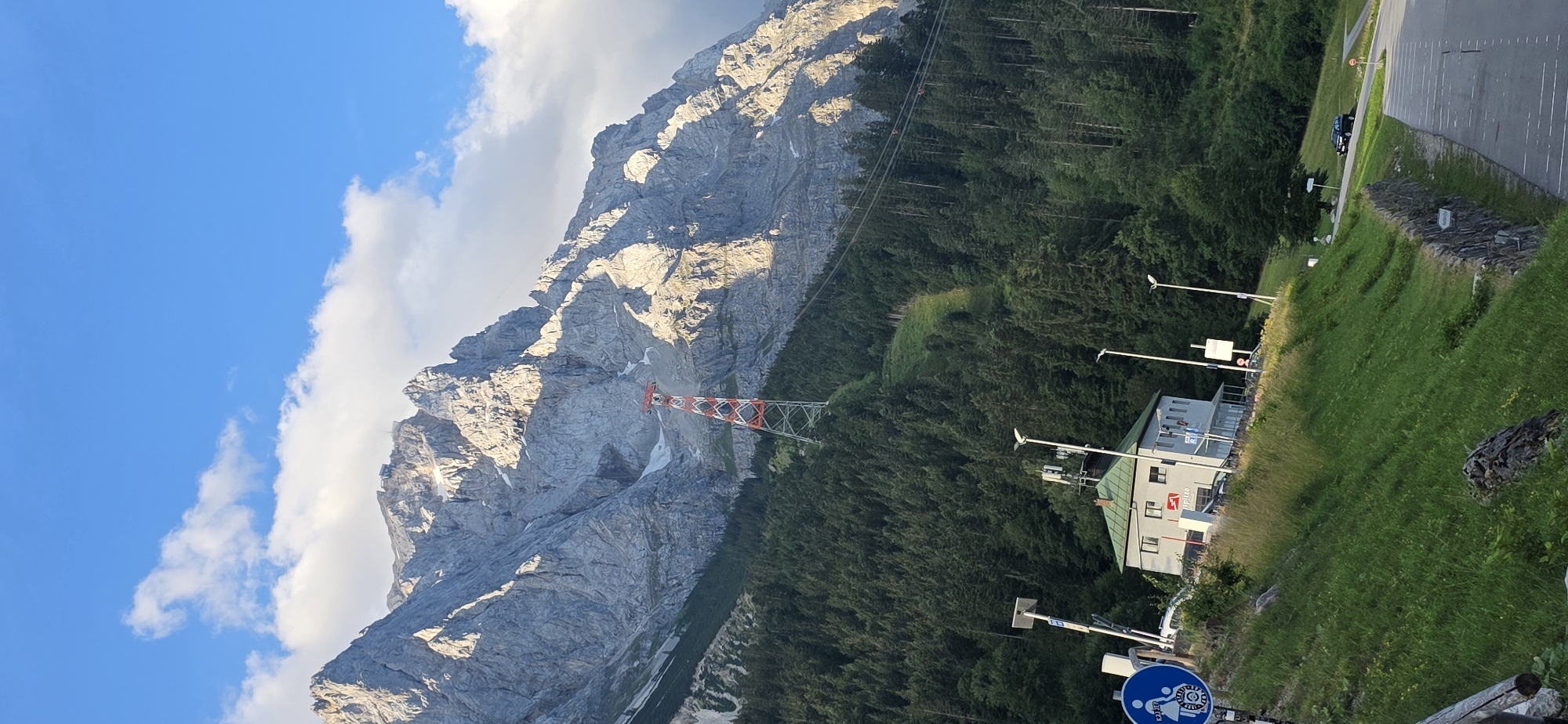 The image depicts the Zugspitze cable car station in Grainau, Germany. In the foreground, a grassy slope is visible, with a paved road and parking area to the right. A stone wall runs along the edge of the slope. On the slope, a small building with a green roof is situated, serving as part of the cable car station. A blue circular sign with a white pictogram of two figures walking is also present.
The middle ground is dominated by a dense forest of evergreen trees, leading up to the imposing Zugspitze mountain range. A red and white pylon for the cable car is prominent against the mountainside, with cables stretching upwards. The mountain itself is rugged and rocky, with patches of snow and sunlight illuminating its contours.
The background shows a clear blue sky with scattered white clouds. The scene is a depiction of a mountainous landscape, typical of the Bavarian Alps, with infrastructure for tourism and recreation. There are no people or animals clearly visible in the scene.