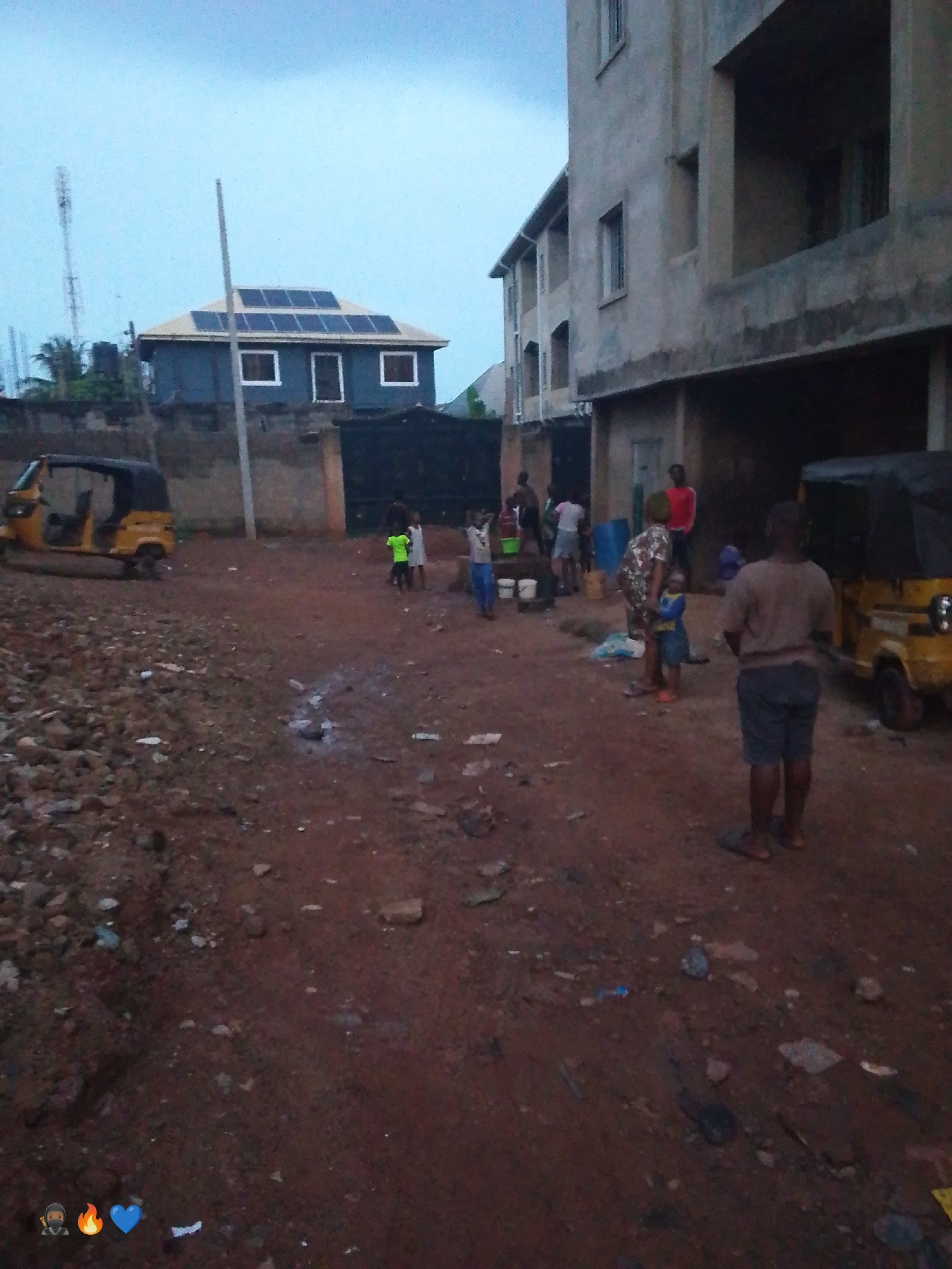 In an outdoor, unpaved area of Unknown, Unknown, several people are gathered near buildings. In the foreground, a person stands facing away from the camera, wearing a grey t-shirt and dark shorts. To the left, a yellow auto-rickshaw is parked. In the mid-ground, a group of children are visible. One child in a green shirt stands near a blue barrel. Further back, a building with solar panels on its roof is visible. Adjacent to it, a multi-story building with unfinished construction is present. Another yellow auto-rickshaw is parked to the right of the frame. The ground is covered in dirt and debris.