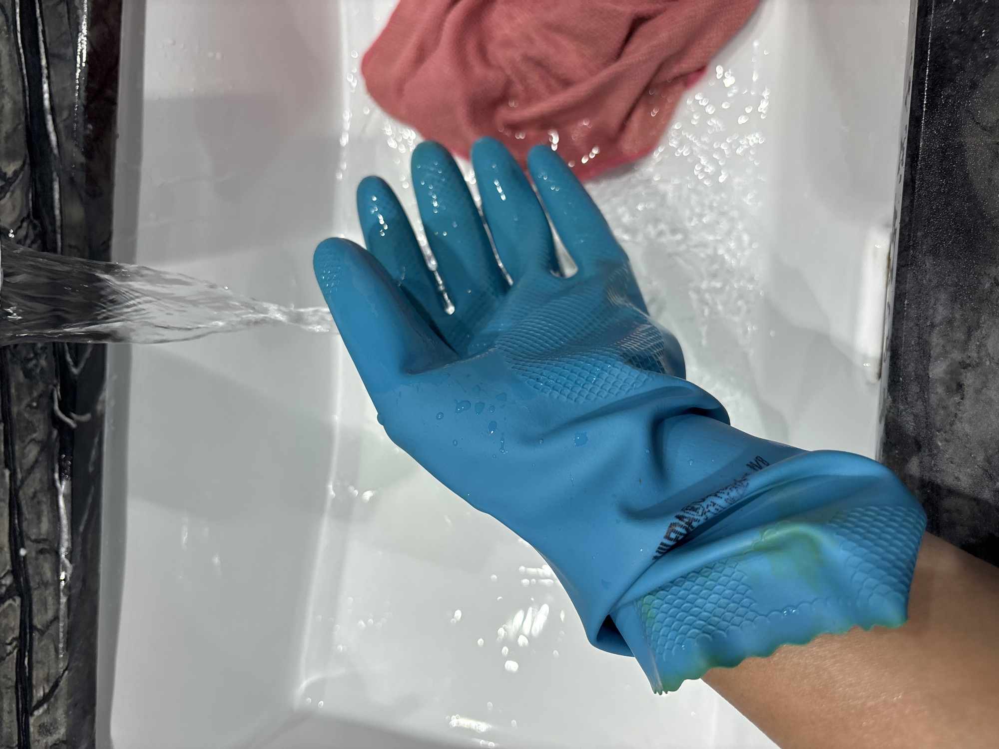 A person's left hand, covered by a blue rubber glove, is held under a stream of water running from a faucet into a white sink. The glove is wet and beaded with water droplets. To the right of the hand and glove, a pink cloth or rag is partially submerged in the sink. The sink appears to be made of porcelain and has a speckled, dark-colored countertop. The background shows the faucet and the top edge of the sink with a textured black material. The scene is indoors, likely in a kitchen or bathroom, and the lighting is bright, suggesting overhead illumination. There is no indication of the location being Dubai, United Arab Emirates.