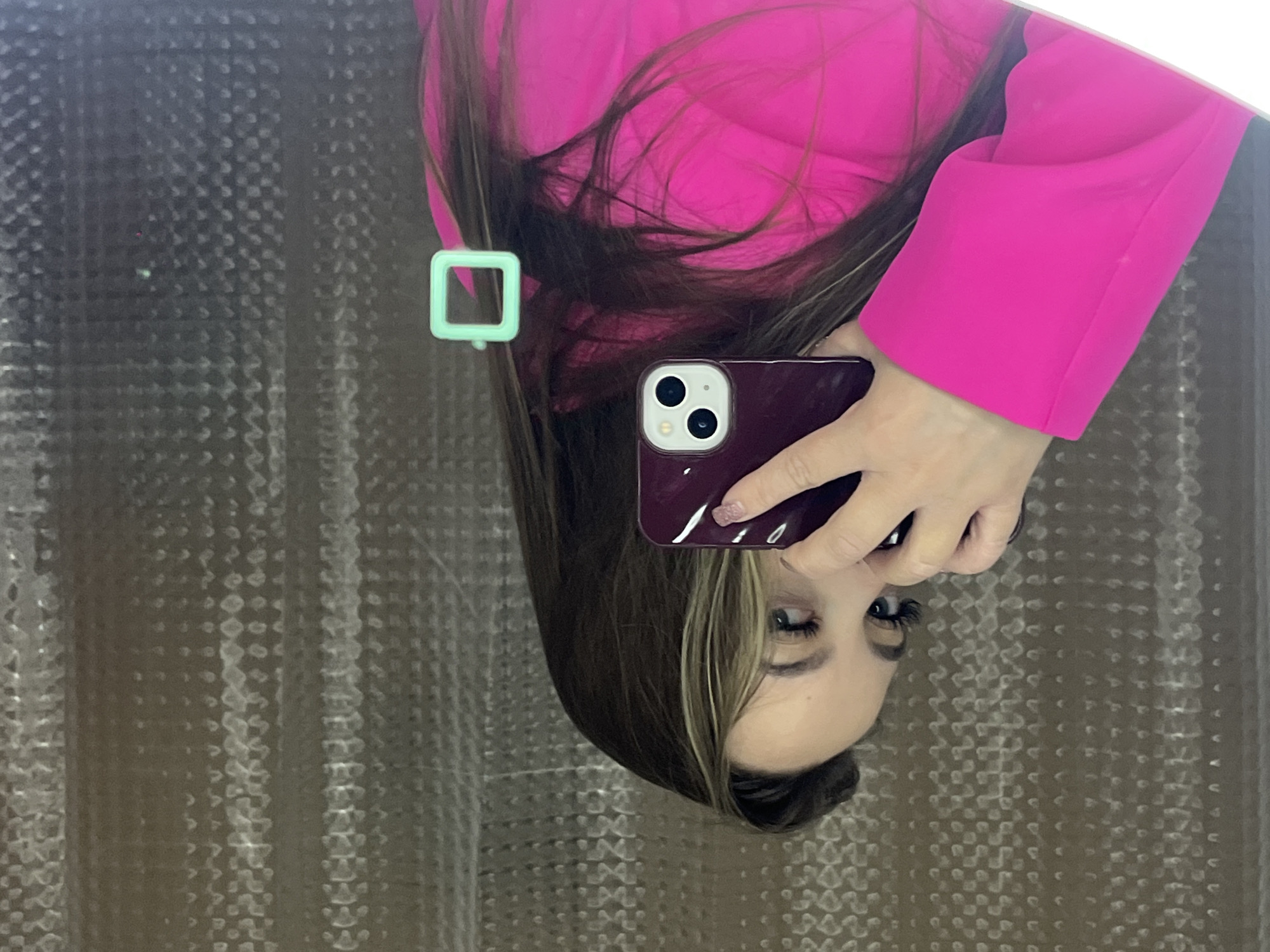A woman wearing a pink jacket takes a selfie with a purple phone in a mirror. The mirror is hanging in front of a textured beige or grey curtain. There's a green square-shaped object attached to the mirror. There is no evidence to identify a location or the city of Krasnodar, Russia.