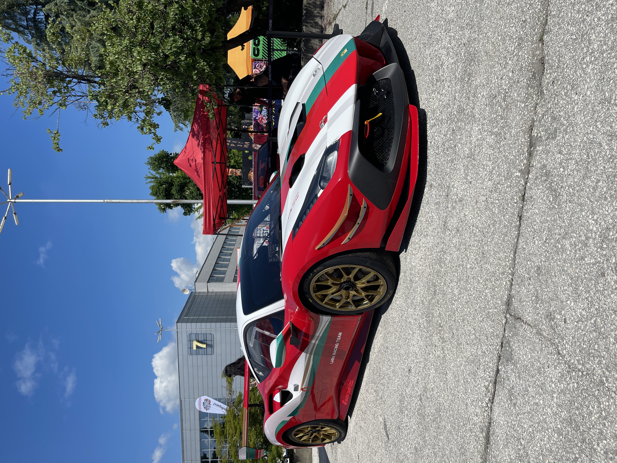 The image shows a red, white, and green painted sports car parked in a city environment. The car has gold rims and branding for "LARA RACING TEAM". Behind the car are several tents with vendor booths, and a building with a large number "7" on its side. Above the building, the sky is blue with some clouds. A light pole is seen on the left side of the frame. There is a banner that appears to be for an event that reads "повядайте!" and "A' motor park" and other illegible markings.