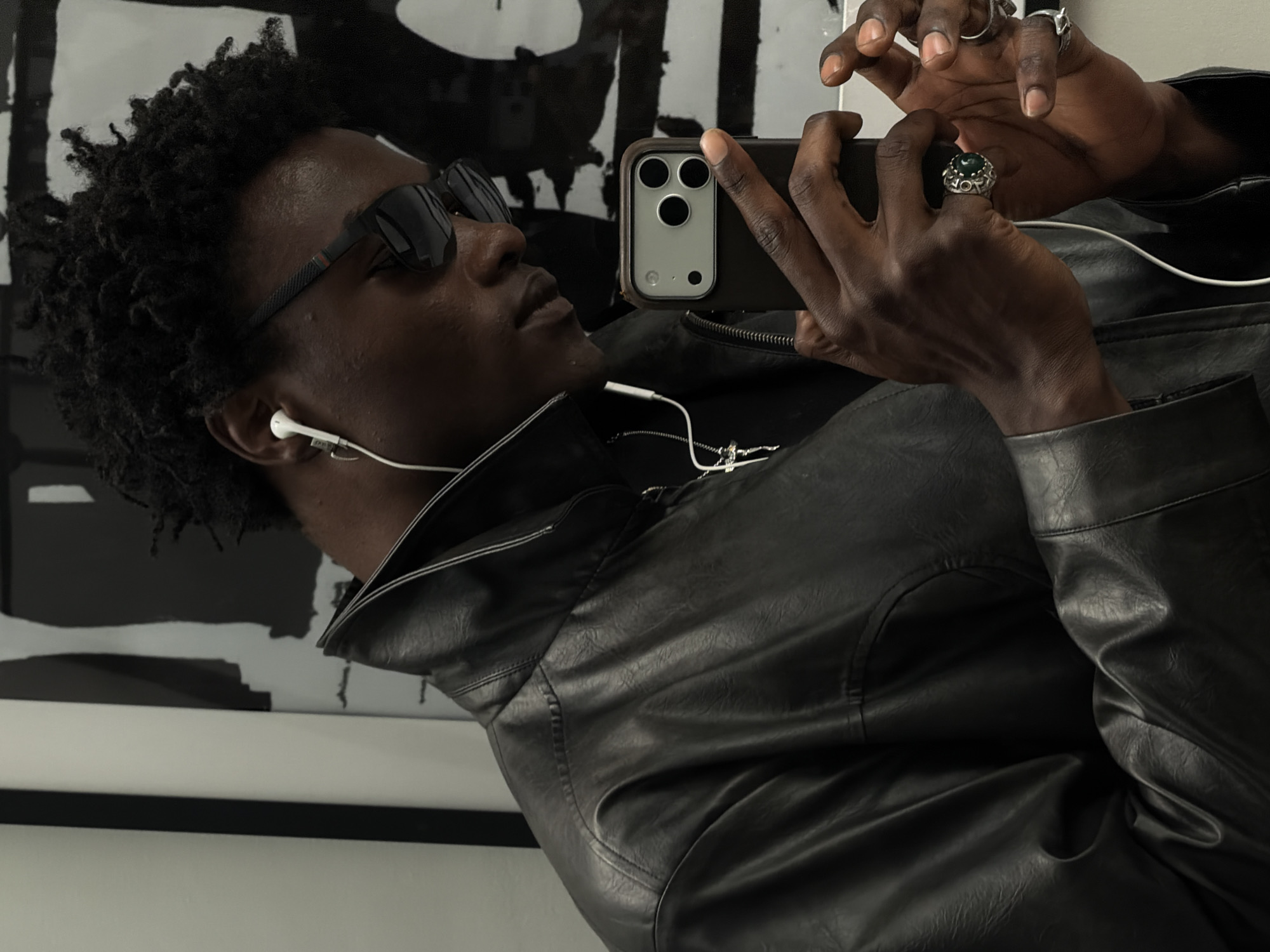 A dark-skinned man with short, curly hair, wearing sunglasses, a black leather jacket and white wired earphones, is holding a phone with multiple lenses. He is taking a selfie with the phone and is wearing several rings on his fingers. There is a framed piece of black and white abstract art behind him, mounted on a white wall. The location is not identifiable as London, United Kingdom.