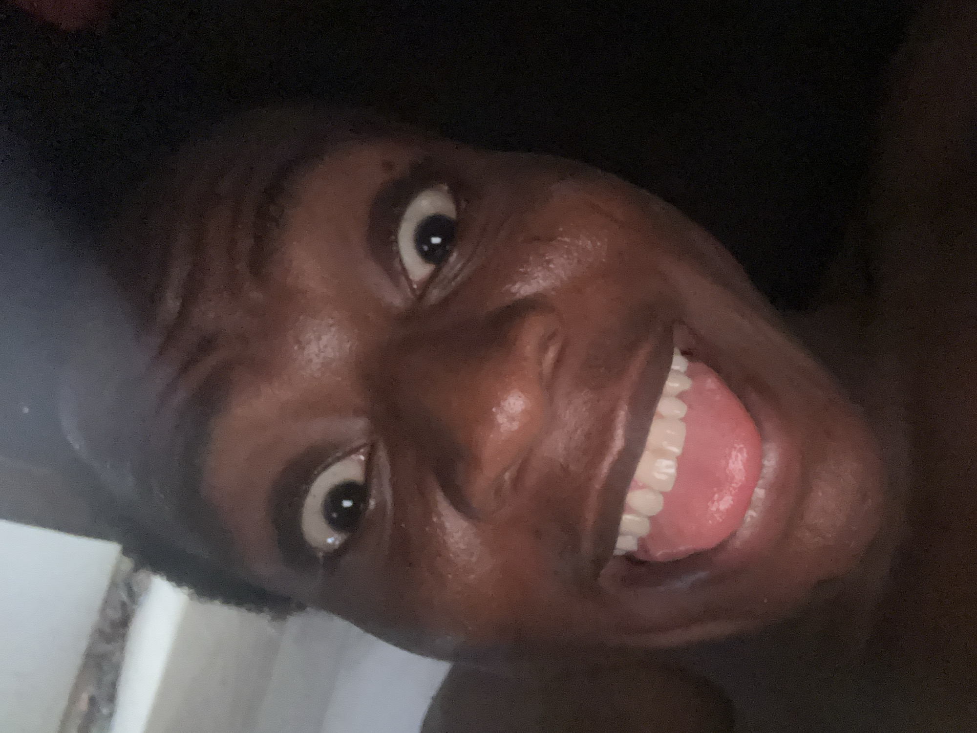 A close-up, slightly angled view of a dark-skinned person's face, with a large smile exposing their teeth and tongue. Their eyes are wide and appear expressive. The background is dark and indistinct. A small section of a white wall or surface is visible in the lower left corner.