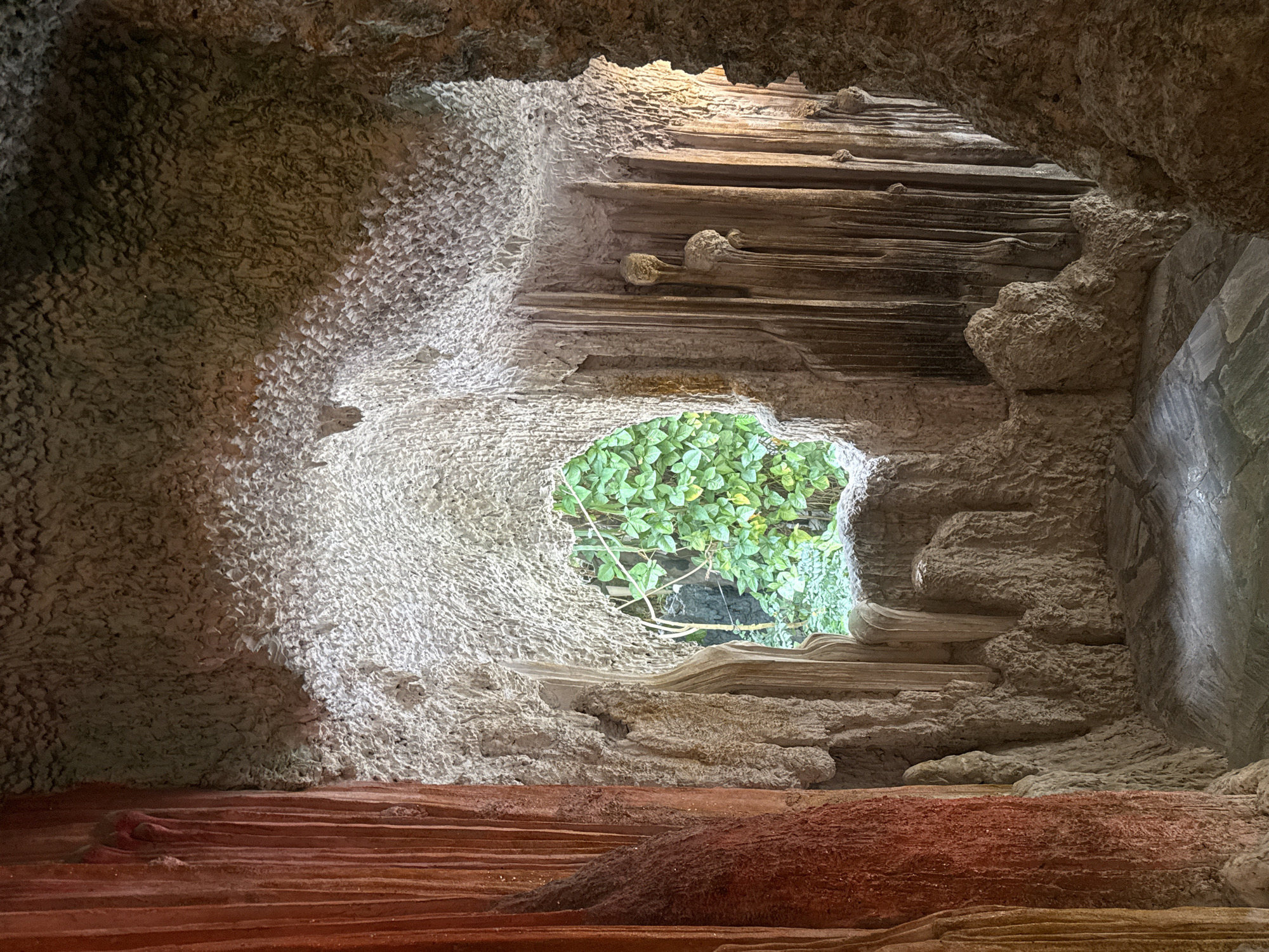 The image depicts an artificial cave-like structure, likely an attraction within a garden or park in Da Lat, Vietnam. The interior features textured walls and stalactite-like formations, sculpted from a material that mimics natural rock. On the left, a vertical element with a reddish-brown, textured surface stands prominently. In the center, an opening reveals a lush green foliage of plants and trees from the outside. The floor appears to be paved with irregular stone tiles. The lighting suggests an indoor or shaded environment, with some areas brightly illuminated and others in shadow. There are no people visible in the image.