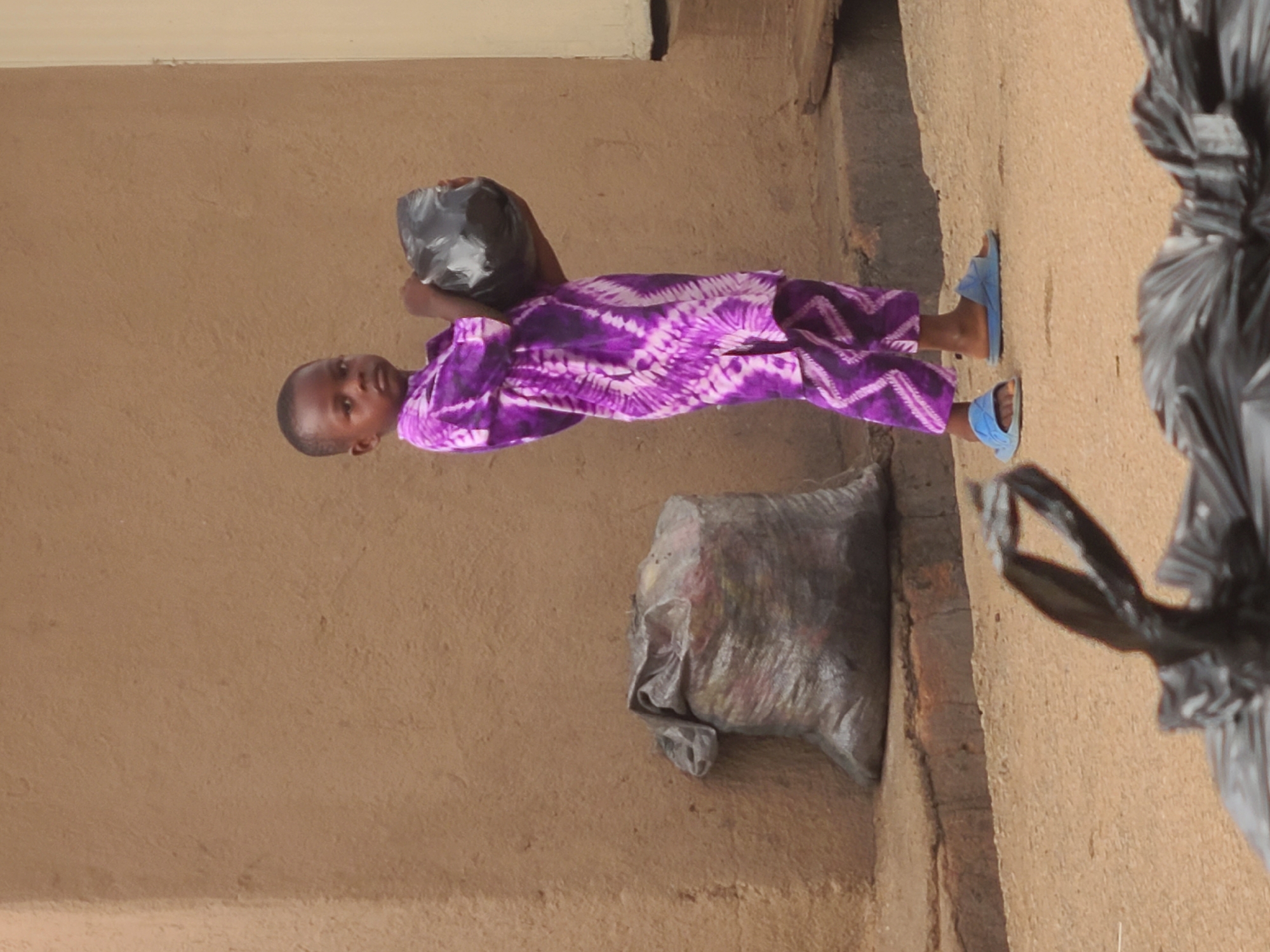 A child wearing a purple and white patterned garment and blue sandals carries a black plastic bag in Jos, Nigeria. The child stands on a dirt-colored ground next to a wall of similar color, with additional bags nearby.