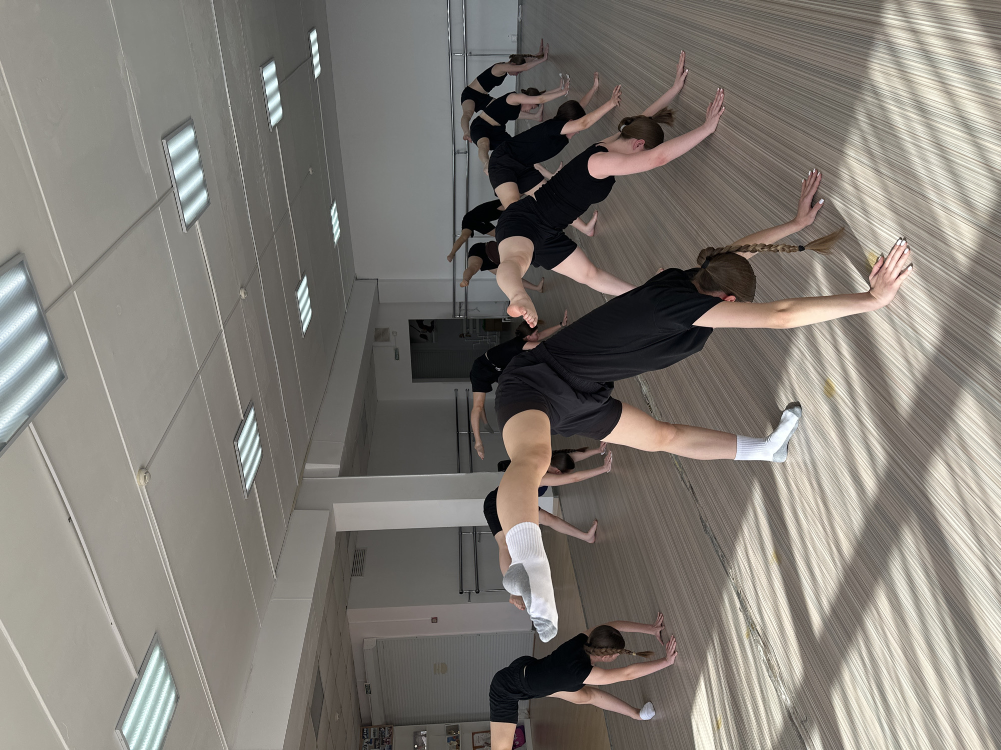 A group of approximately ten young women dressed in black athletic wear are engaged in a stretching exercise in what appears to be a dance or exercise studio. The women are aligned in a row, stretching towards a wall. Some of them are wearing socks. The studio has a wooden floor and a white ceiling with recessed fluorescent lighting. There are ballet barres along the walls. The location is likely a dance or gymnastics studio in Votkinsk, Russia, based on the image's origin.
