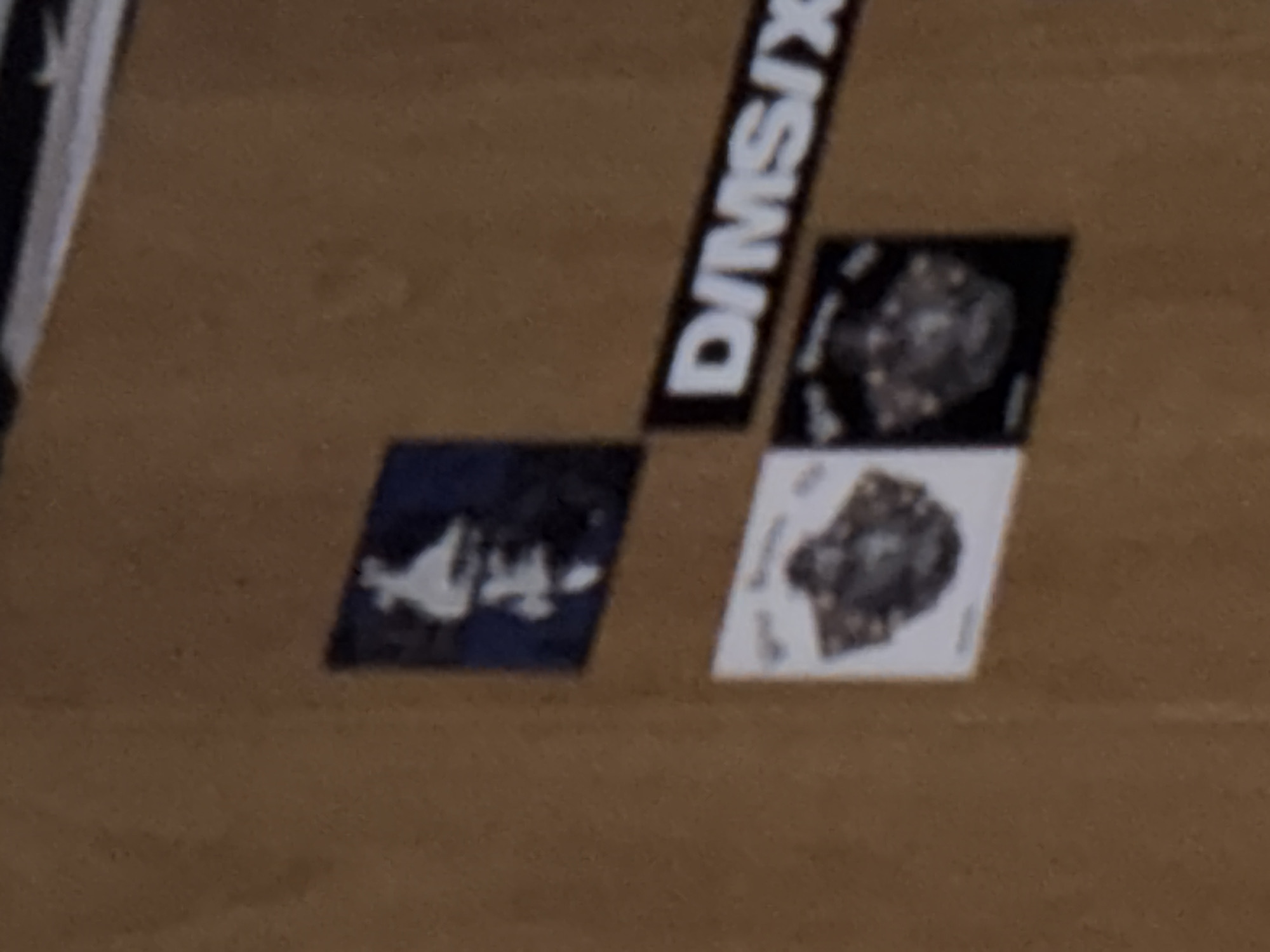 A close-up, slightly blurry image displays several stickers affixed to a warm brown, textured surface, possibly wood or concrete. A black rectangular sticker bearing the white capital letters "DIMS IX" is prominently positioned diagonally across the upper central part of the frame, running from the top-right to the bottom-left.
To the left and slightly below the "DIMS IX" sticker is a dark blue square sticker. This blue sticker features a light-colored, somewhat indistinct graphic that appears to be an abstract symbol or a stylized figure.
Further below the diagonal sticker and to its right, two more square stickers are arranged. The top-right of these two is black and showcases a dark, intricate graphic, which appears to be a highly detailed skull or a complex mechanical component, rendered in shades of grey. Adjacent to it, on the bottom-left, is a white square sticker. This white sticker displays a dark and intricate graphic very similar to, if not identical with, the one on the black sticker, also resembling a skull or mechanical part. The fine details of the graphics on the square stickers are present but slightly softened by the overall blurriness of the image. The background surface exhibits subtle variations in color and texture. No people or activities are visible.