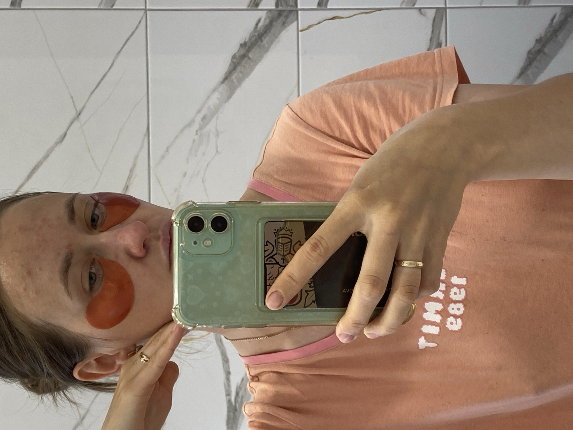 A young woman with fair skin and auburn hair pulled back in a messy bun, wearing an orange t-shirt with pink trim, takes a mirror selfie in a bathroom. She has red under-eye patches on her face, and is wearing two gold rings on her left hand. She is holding a mint-green iPhone with a heart pattern on the case. The background shows a white tiled wall with gray marble accents.