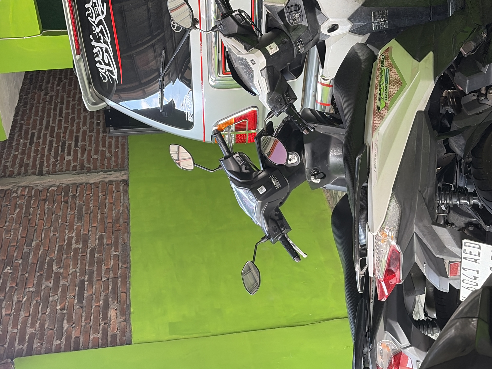 In Boja, Indonesia, a close-up shot focuses on the rear ends of two parked scooters, a silver van, and a brick wall. The scooters are positioned side-by-side, with the majority of their backs visible. The scooter on the right is predominantly white and gray, while the one on the left is black and gray. Both scooters feature prominent red taillights and what appear to be license plates. The van, positioned behind the scooters, is silver and has a sticker with white Arabic script on its rear window. The background is dominated by a vibrant green wall, contrasting with the rough texture of the red brick wall above it. The lighting suggests it is daytime, with shadows indicating sunlight from the right.