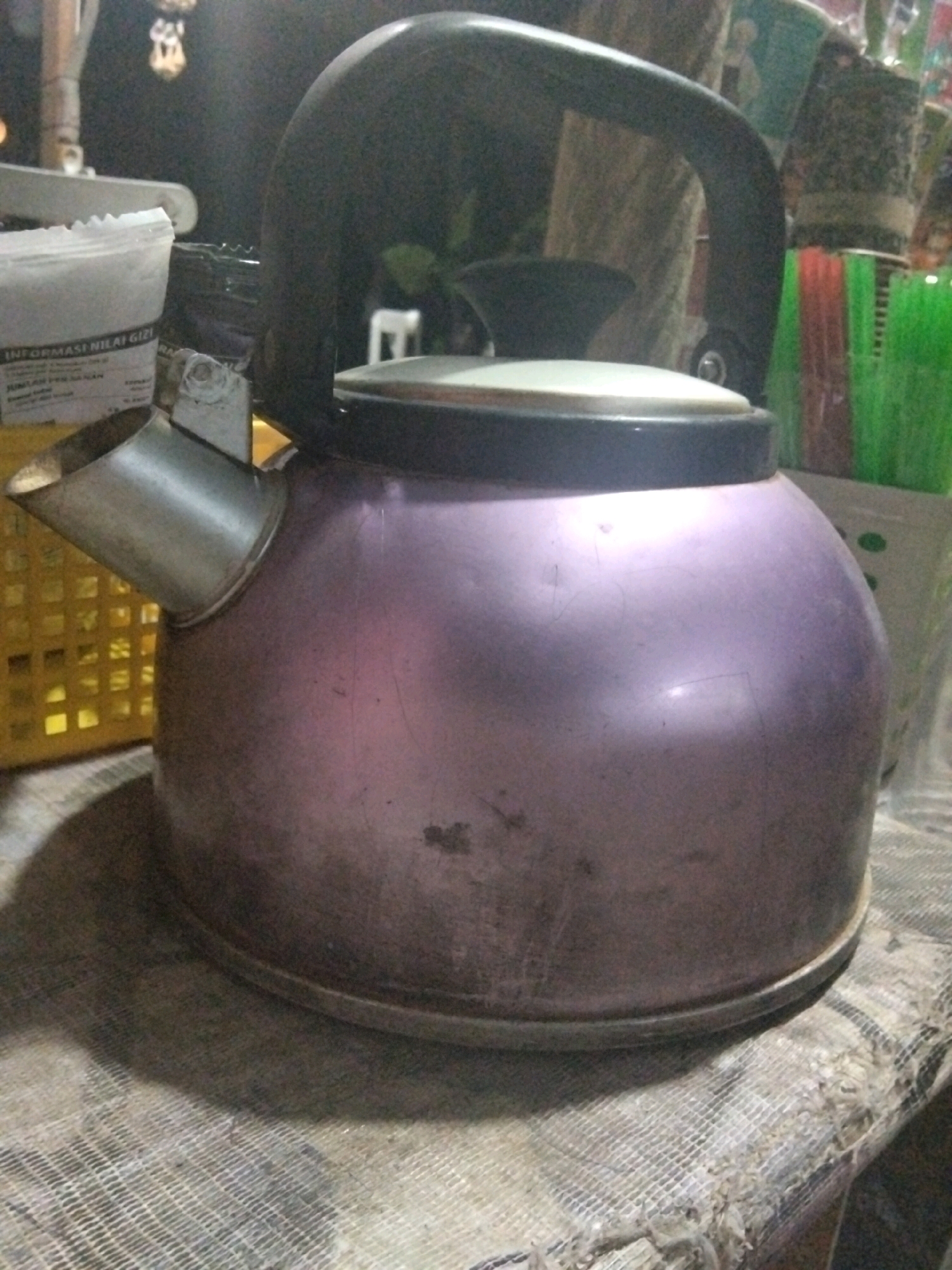 A purple kettle with a black handle and a metallic spout sits on a textured surface. The kettle is dusty, with visible scratches. Behind the kettle, a yellow plastic basket is partially visible, containing what appears to be a bag of white material with Indonesian text, possibly nutritional information. Further in the background, a collection of colorful brushes, predominantly green and red, are arranged in a container. The lighting is dim, casting shadows and creating a somewhat hazy atmosphere. The scene appears to be an indoor setting, possibly a kitchen or storage area, within a residential context in Cikal, Indonesia. There are no individuals present in the image.