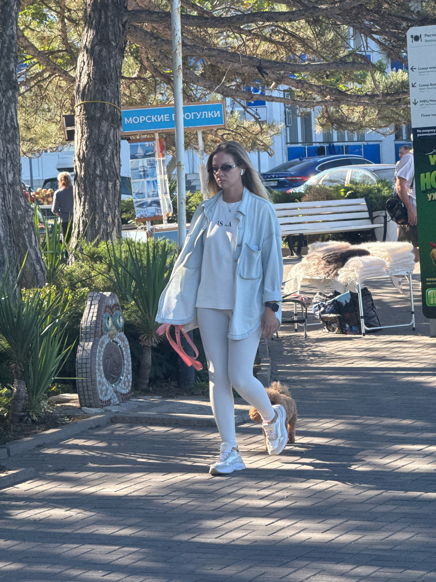 A woman wearing a light-colored top and leggings, white sneakers, and sunglasses is walking a small, tan dog on a leash in the city of Anapa, Russia. She has earbuds in her ears. The scene takes place on a paved walkway, with trees and lush green foliage lining the path. In the background, there are several buildings, including one with a sign that appears to be in Russian. A bench and a table with stacked items are also visible. The lighting suggests it is daytime, with shadows cast on the walkway.