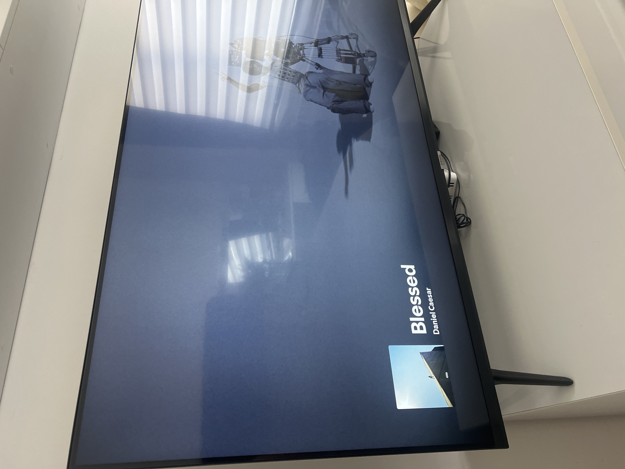 A television displays the album art for "Blessed" by Daniel Caesar. The television is black with a stand on the right. The wall and a window with blinds are behind the television. The location is Unknown, Unknown.