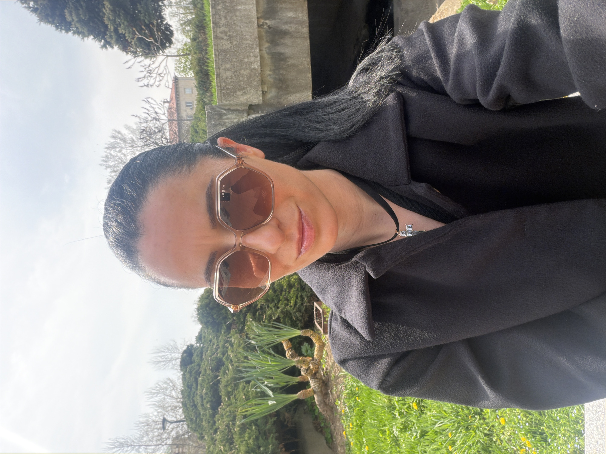 A woman with dark hair, wearing rose-tinted sunglasses and a dark overcoat, poses for a selfie. The background consists of trees, green grass, and a concrete wall, suggesting an outdoor setting in a park or garden. A building is partially visible in the distance. Considering the user's request for the location in Pleven, Bulgaria, the background elements are consistent with the landscape found in Pleven's city parks and gardens.