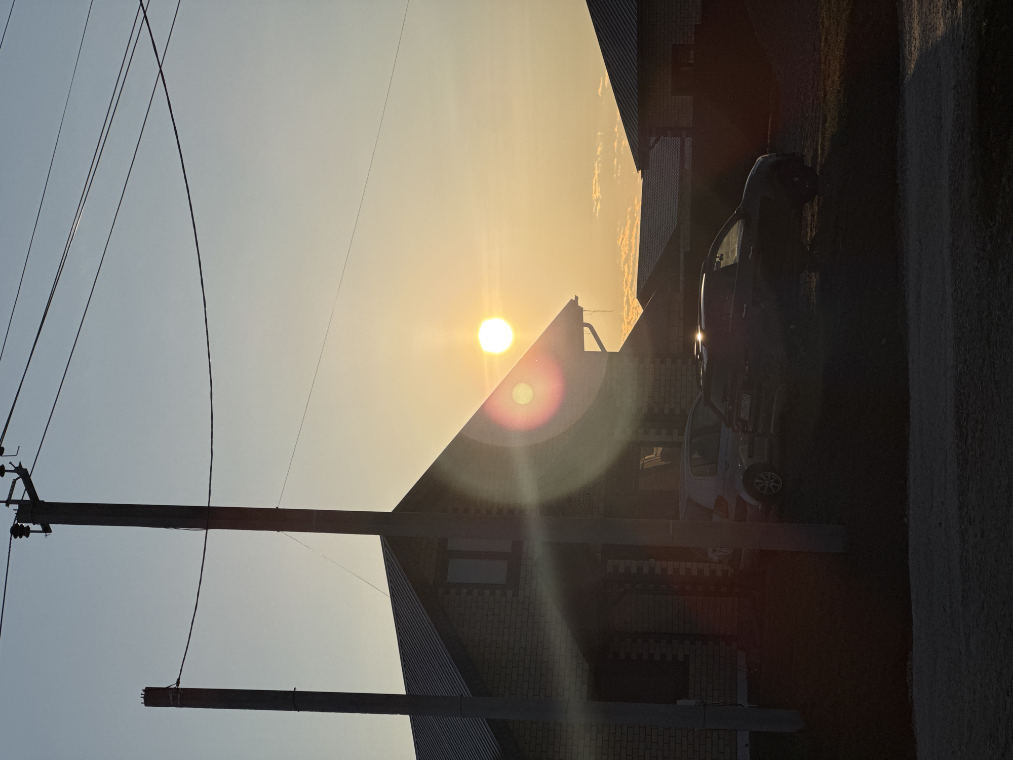 A street scene in Ферма, Russia shows a brick house silhouetted against a bright sun. Power lines and a utility pole are in the foreground. Two cars are parked along the side of the road, obscured by shadows. The street is asphalt. The image is rotated to the side.