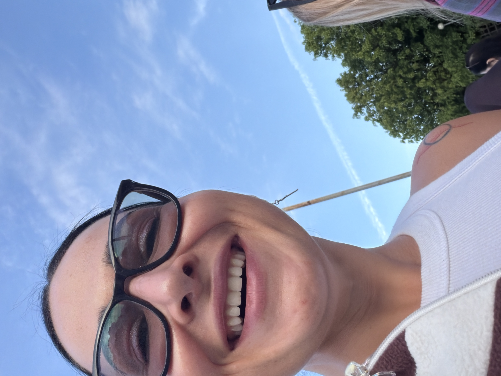 A selfie-style image captures a person smiling broadly, positioned against a clear, bright blue sky in Saint Petersburg, Russia. The individual, likely female, is looking directly at the camera from a low angle, showing their face in the lower left portion of the frame. They are wearing black-framed glasses, and their mouth is open in a wide, joyful smile, revealing white teeth. Their dark hair is pulled back.
Behind the smiling individual, a thin, light-colored pole or stick extends upwards. To the right, a white tank top or undershirt is visible, and below it, a brown and white striped garment with a zipper. A tattoo, appearing as a red and black circular design with radiating lines, is visible on the person's upper arm/shoulder. In the upper right corner, a portion of blonde hair and the top of another person's head are visible, alongside lush green tree foliage.
The background features a vibrant blue sky with scattered wispy clouds and a prominent, long, white contrail from an aircraft, stretching diagonally across the frame. The lighting suggests it is daytime, likely midday or afternoon, under clear and pleasant weather conditions. No text is visible in the image.