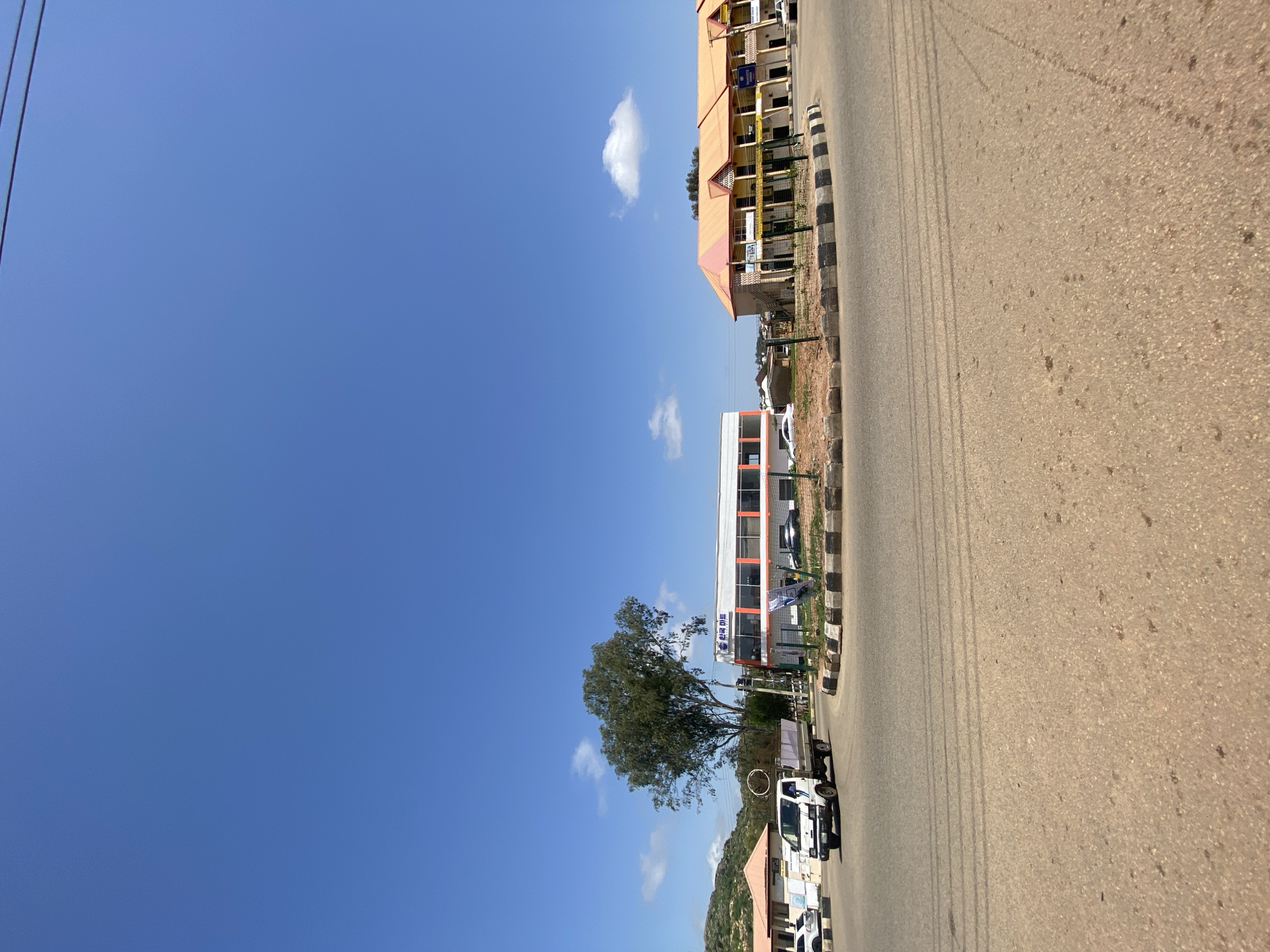 The image captures a street scene in Gura Top, Nigeria, under a clear blue sky with scattered clouds. In the foreground, a paved road curves around a traffic island adorned with black and white checkered borders. A white truck with a gray cargo bed is visible, navigating the road. To the left of the traffic island, a building with an orange and white facade and "한국마트" (Korean Mart) signage is present. Across the street, a longer, multi-story building with an orange roof and yellow exterior, featuring balconies and shops on the ground floor, is visible. Further in the background, a large, leafy tree stands prominently. The environment appears to be a commercial or mixed-use area in a developing urban setting.
