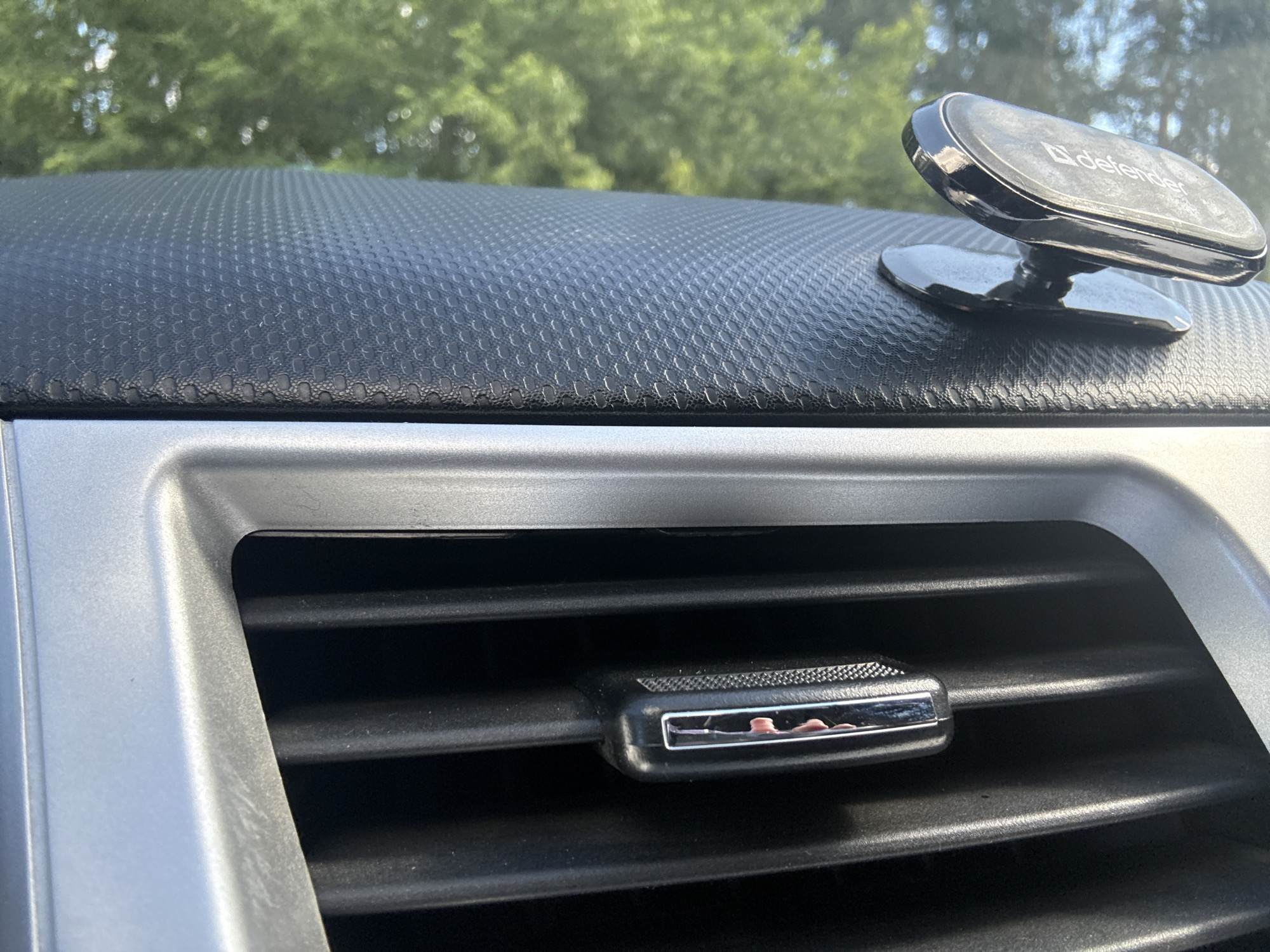 The image captures a close-up view of a car's interior, focusing on the dashboard and an air vent. The dashboard has a textured, dark grey surface with a honeycomb-like pattern. A black magnetic phone mount, branded "defender", is attached to the dashboard on the right side of the frame. The mount has a sleek, glossy finish. In the lower portion of the image, a silver-colored trim surrounds a black air conditioning vent. A small, rectangular air freshener or clip is attached to one of the vent slats, featuring a chrome edge and a patterned surface. Inside the air freshener, a faint pinkish-red substance is visible, possibly indicating its scent. The background of the image is out of focus, showing green foliage, suggesting that the car is outdoors, possibly in a natural setting or a wooded area. The lighting indicates it is daytime, with the sun casting highlights on the car's interior surfaces. There are no people visible in the image. The setting is an automobile interior, likely parked. The overall impression is one of a functional and somewhat personalized car interior.