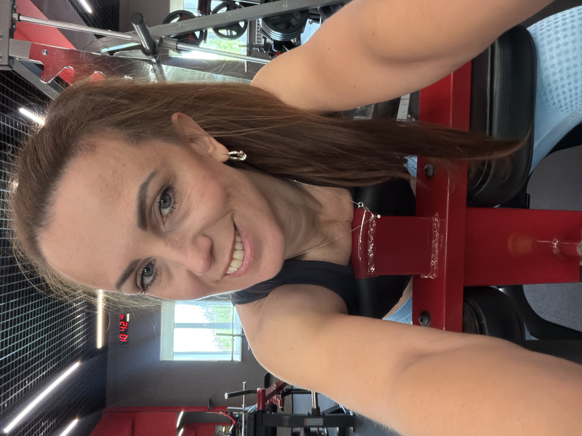 A selfie is taken by a woman in a gym. She has long brown hair, blue eyes, and is smiling. She is wearing a black tank top and blue leggings. She is leaning on a red gym machine. In the background, there are gym weights and a clock on the wall that reads 10:42. The gym appears to be in Birobidzhan, Russia.