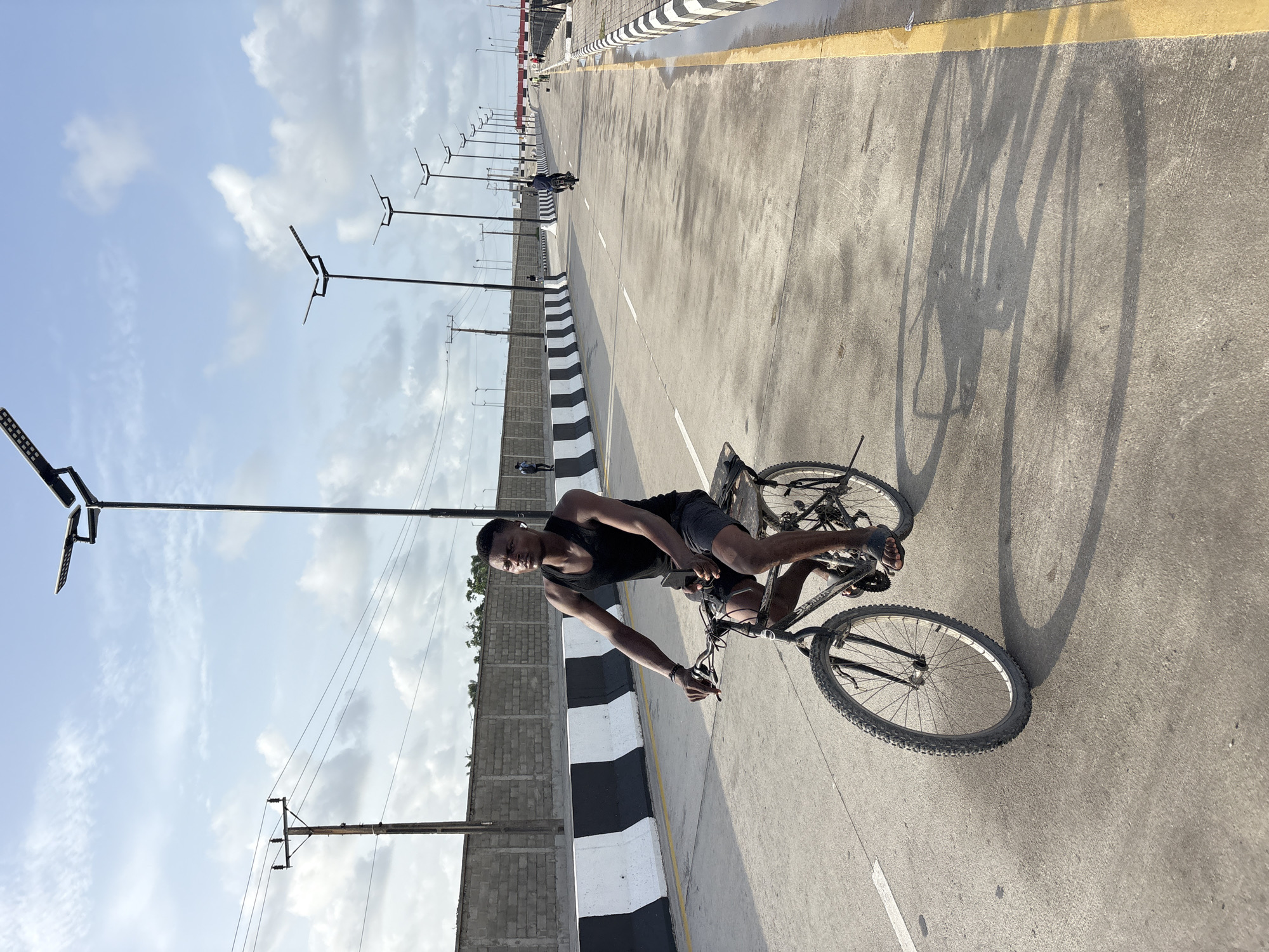 An African man is riding a bicycle on a paved road. The road is divided with black and white striped barriers. A concrete wall runs alongside the road with lamp posts attached to it. Power lines are visible. In the distance, another person on a bicycle is also on the road. The sky is cloudy with blue patches. The location is Igboefon, Nigeria.