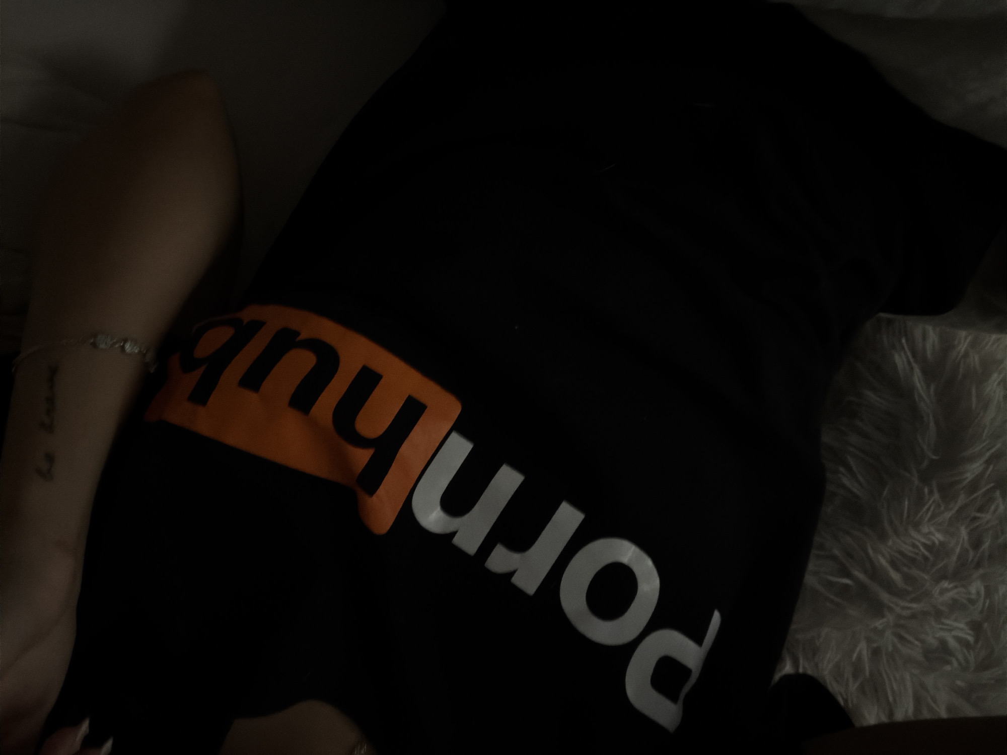 A person is lying on what appears to be a white, fluffy surface, possibly a bed or a rug. The person is wearing a black t-shirt with the "Pornhub" logo printed in white and orange. Part of the person's arm is visible on the left, adorned with a silver bracelet and a tattoo. The lighting in the image is dim, making it difficult to discern further details. There is no indication of Sofia, Bulgaria, in the image.