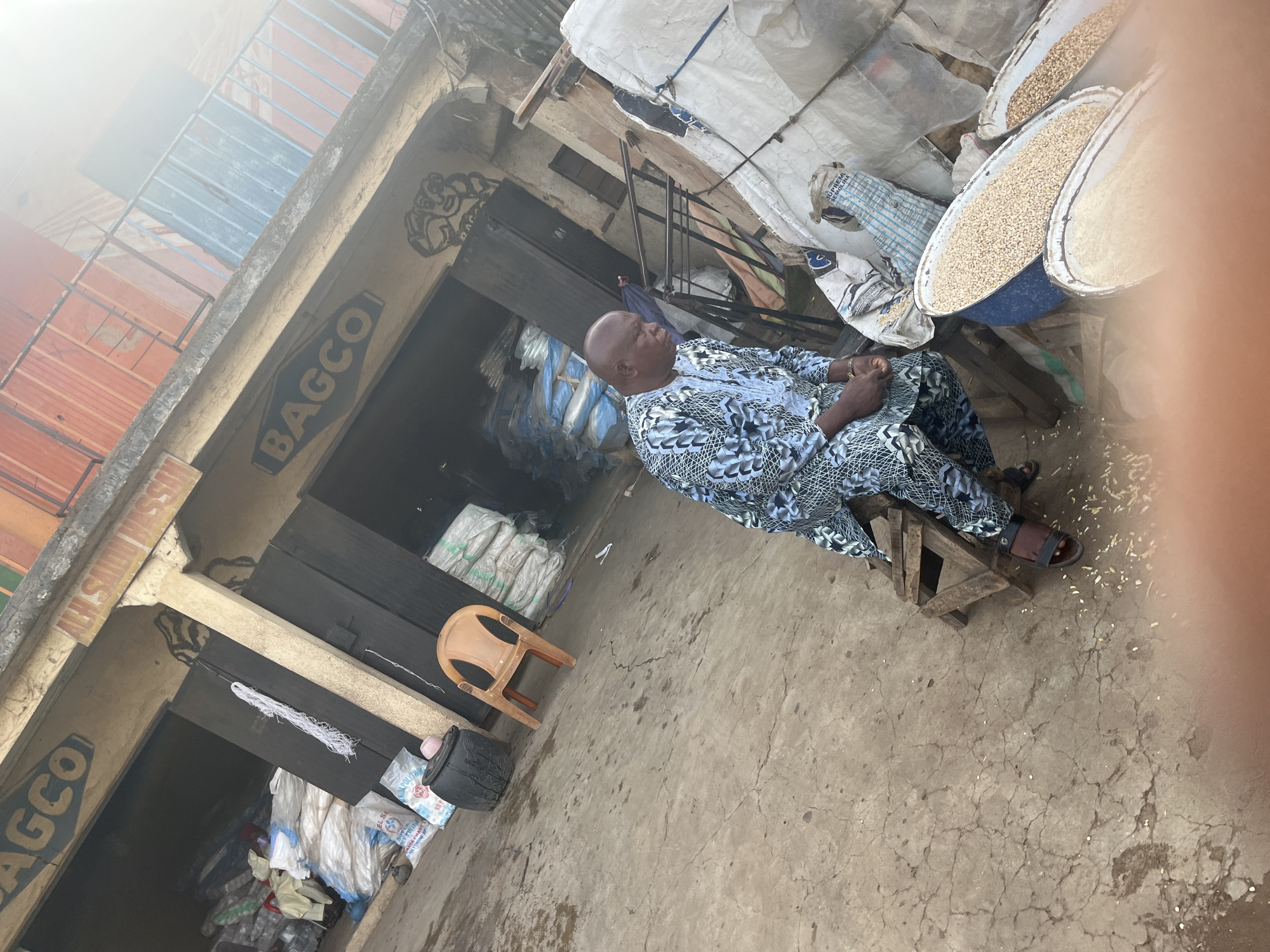 In Jos, Nigeria, a man is seated on a wooden stool outside a shop. He is wearing a light blue and white patterned traditional African garment and dark sandals. The shop, identified by a sign reading "BAGCO," appears to be a market stall or warehouse. Bags of what appears to be grain or produce are stacked outside, and in the foreground, there are three large shallow baskets or trays filled with similar material. The ground is concrete and cracked, with scattered debris. The overall scene depicts a commercial setting in an outdoor market environment.