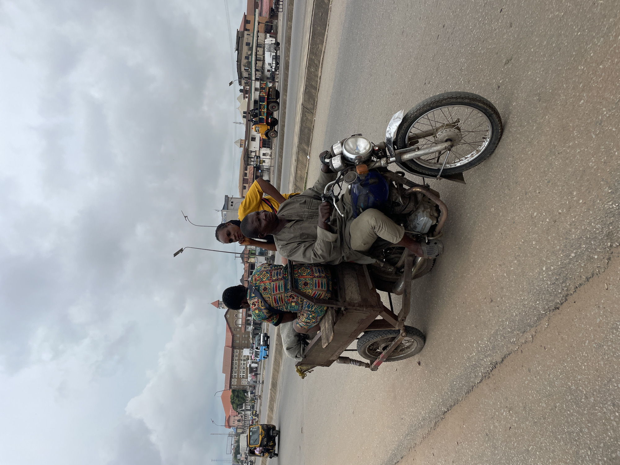 The image shows a man riding a motorcycle with a trailer in an urban environment, possibly in Africa, given the presence of a yellow motorized rickshaw and the general architecture. The motorcycle is pulling a wooden trailer with two passengers. The driver is wearing a khaki shirt and pants, and the passengers are a woman in a colorful patterned dress and another woman in a yellow shirt. The trailer appears to be carrying some cargo, possibly sacks. The road is paved, and there are other vehicles and buildings in the background, suggesting it is a commercial or busy area. The sky is overcast, indicating a cloudy day.
