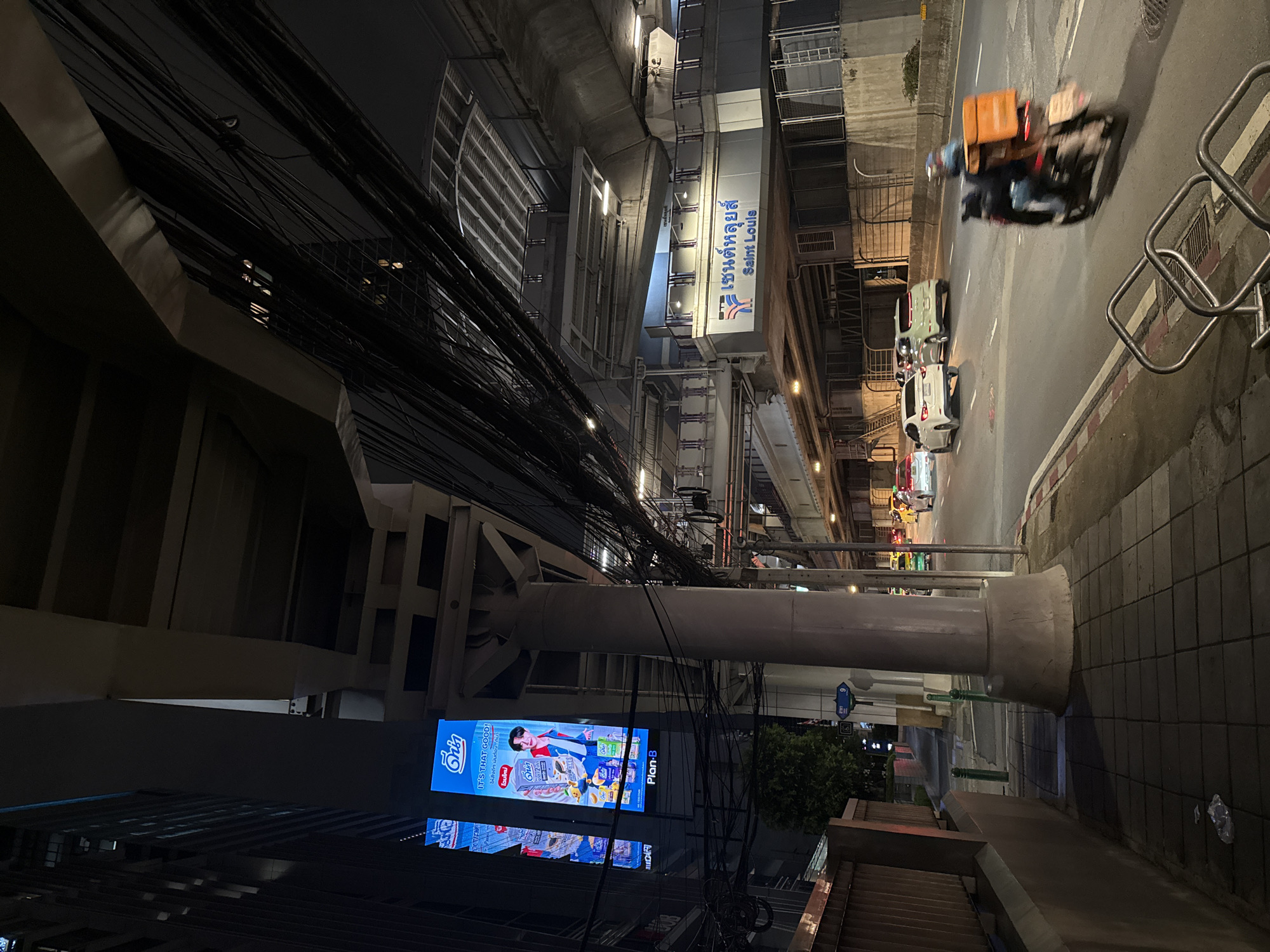 This is a nighttime, low-angle shot of a street scene in Bangkok, Thailand. The image is dominated by a complex network of black electrical wires overhead, casting shadows and obscuring much of the background buildings.
In the mid-ground, a section of an elevated structure, likely a train or skywalk, is visible. It has a modern, gray concrete design with illuminated panels and a sign in Thai and English that reads "เซนต์หลุยส์ Saint Louis." Below this, an illuminated billboard displays an advertisement for a product called "ดีน่า" (Dina), featuring a smiling young man and various products. The text on the billboard includes "IT'S THAT GOOD!" and "Plan-B."
On the street below, several vehicles are visible, including a blurred orange and black three-wheeled vehicle with a passenger, and a few cars. The street is paved with asphalt and bordered by a sidewalk with square paving stones. The overall impression is of a busy urban environment at night, with a mix of infrastructure, advertising, and traffic. The time of day is clearly night due to the artificial lighting and the dark sky.