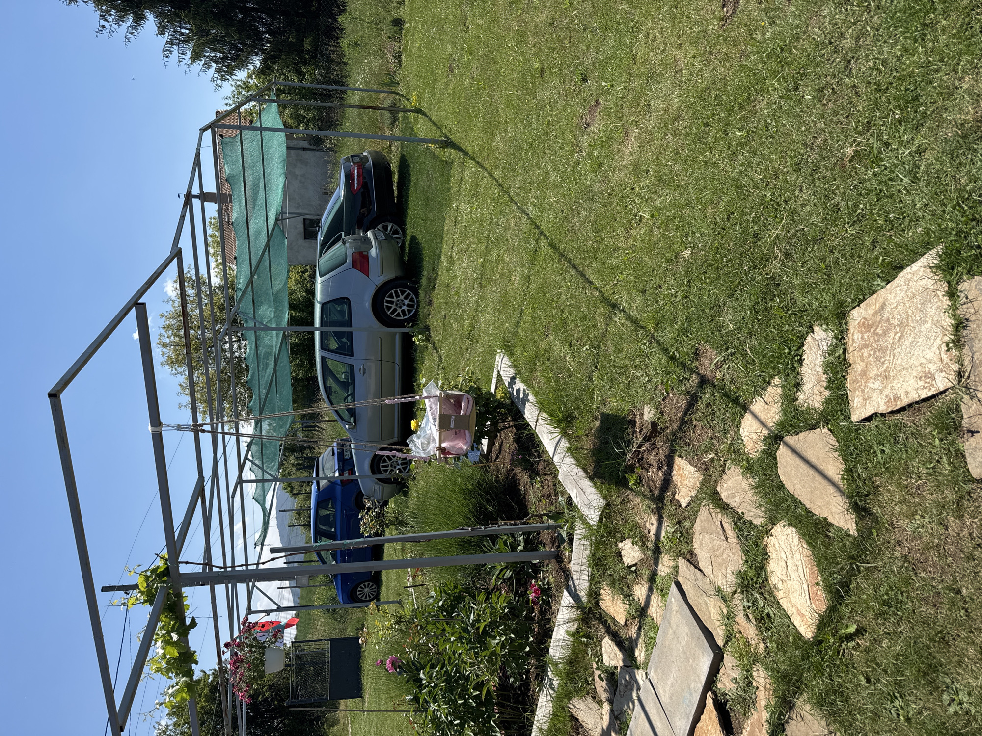 This image depicts a residential garden area in Dolna Dikanya, Bulgaria, under a clear blue sky on a sunny day. A metal pergola, constructed from square tubing, occupies the central and upper frame. A grapevine with green leaves grows along its top-left section. A white hanging planter containing red-pink flowers is suspended from the pergola on the left. A pink plastic child's swing, wrapped in clear plastic, hangs from the pergola on the right, positioned over a raised garden bed.
The raised garden bed, edged with concrete or wood, contains various green plants, including pink peonies and lavender-like bushes. A path of irregular flat stone pavers is laid across the grass in the foreground. Three vehicles are parked under a section of the pergola partially covered with green shade netting: a blue Skoda Fabia hatchback, a silver Volkswagen Golf Mk4 hatchback, and a dark Mercedes-Benz E-Class sedan.
To the left, a dark green metal gate is visible. A white and red flag or textile is affixed near this gate. In the background, a small, light-colored building with a red-tiled roof and a chimney is visible through trees on the right. Distant green hills are present on the horizon under the clear sky. A tall conifer tree stands to the far right. Sunlight produces distinct shadows from the pergola structure and vegetation across the lawn. No people are visible in the scene.