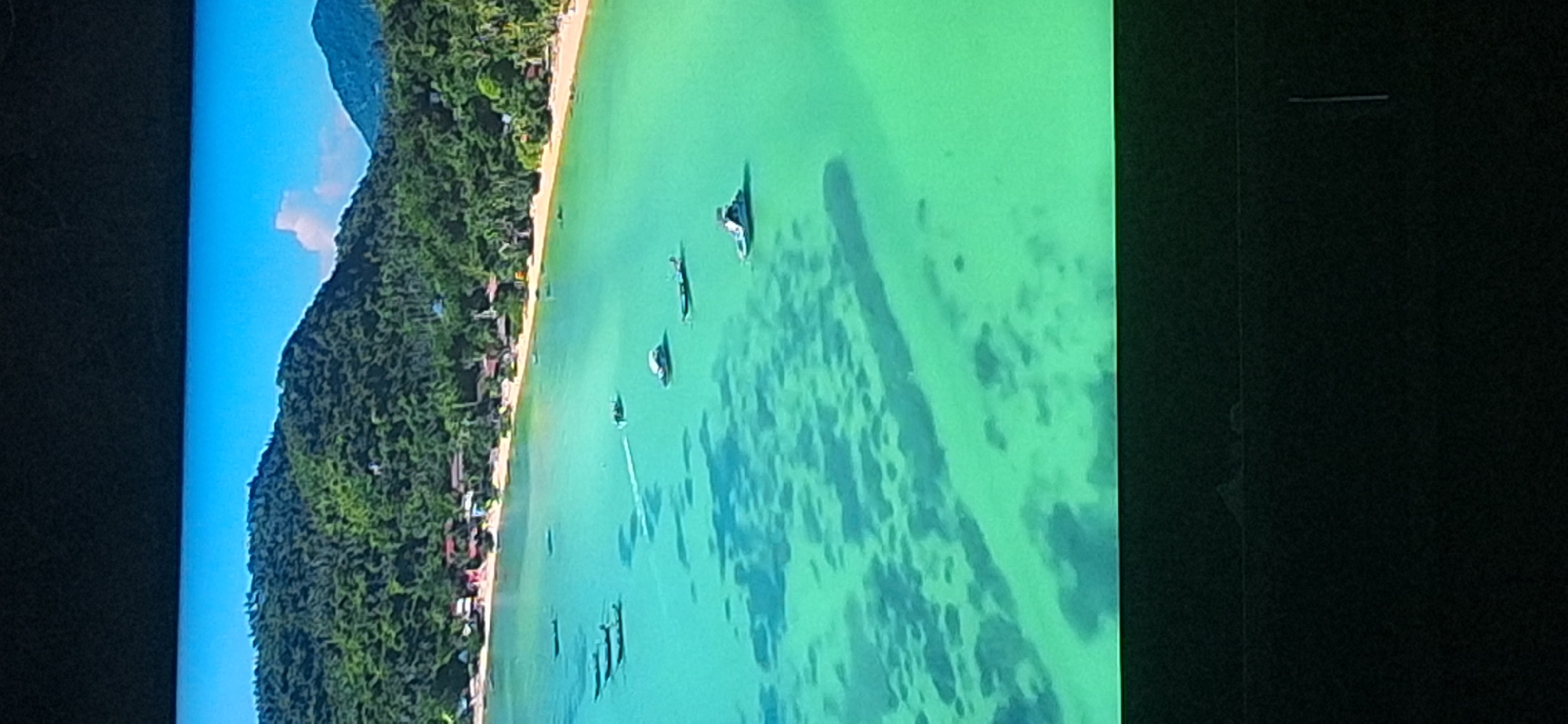 This image shows an aerial view of a tropical coastline with clear turquoise water. There are several small boats scattered in the water, and a sandy beach lined with lush green trees and some buildings. The water is shallow enough to see the sandy bottom in places, with some darker patches possibly indicating coral or submerged rocks. The sky is a bright blue with a few white clouds. The image is a still from a video, as indicated by the slight motion blur on one of the boats and the bright line across the top. The location is NOT in Bila Tserkva, Ukraine, as Bila Tserkva is a landlocked city.