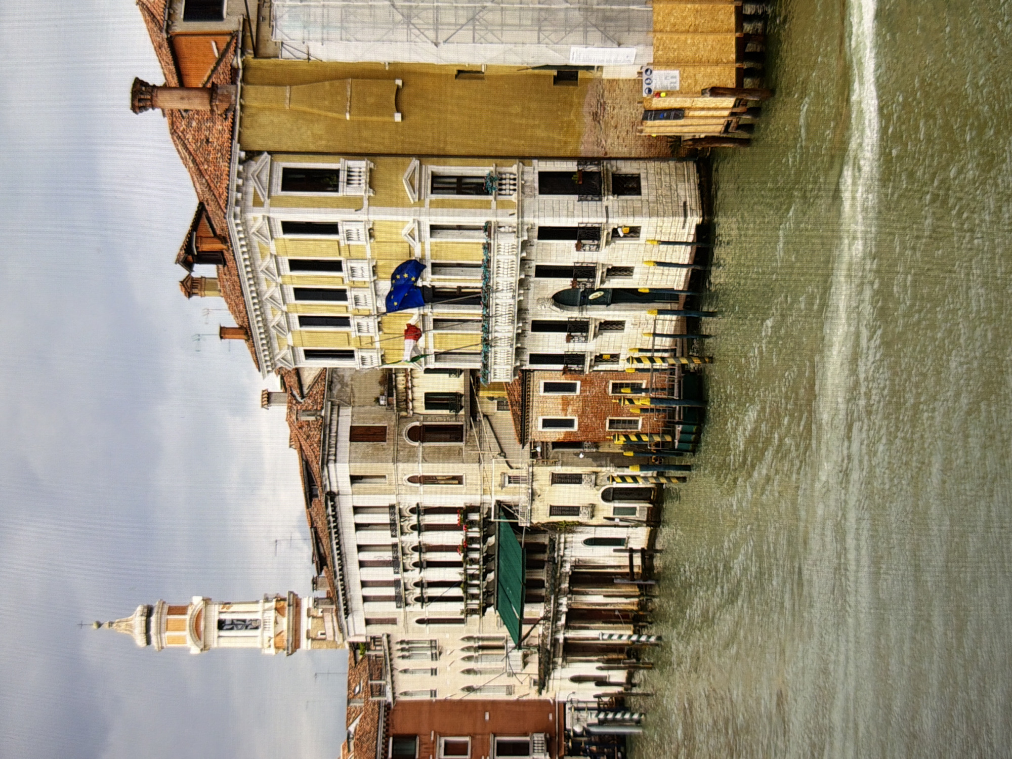 The image depicts a canal in Venice, Italy, with buildings lining its banks. The buildings are old, with terracotta roofs and weathered facades. They are of varying heights and architectural styles, with some featuring ornate details like balconies and arched windows. A tall bell tower with a white and orange spire rises above the buildings on the left side of the image. The canal itself is a murky green, with ripples and waves suggesting movement. Wooden posts, some striped with yellow and black, mark the edge of the canal and appear to serve as mooring points. In the foreground, the water occupies the lower half of the frame, showing the texture of the rippling surface. The sky is overcast with clouds, casting a subdued light on the scene. The overall impression is one of a historic cityscape, with the architecture reflecting centuries of history.
