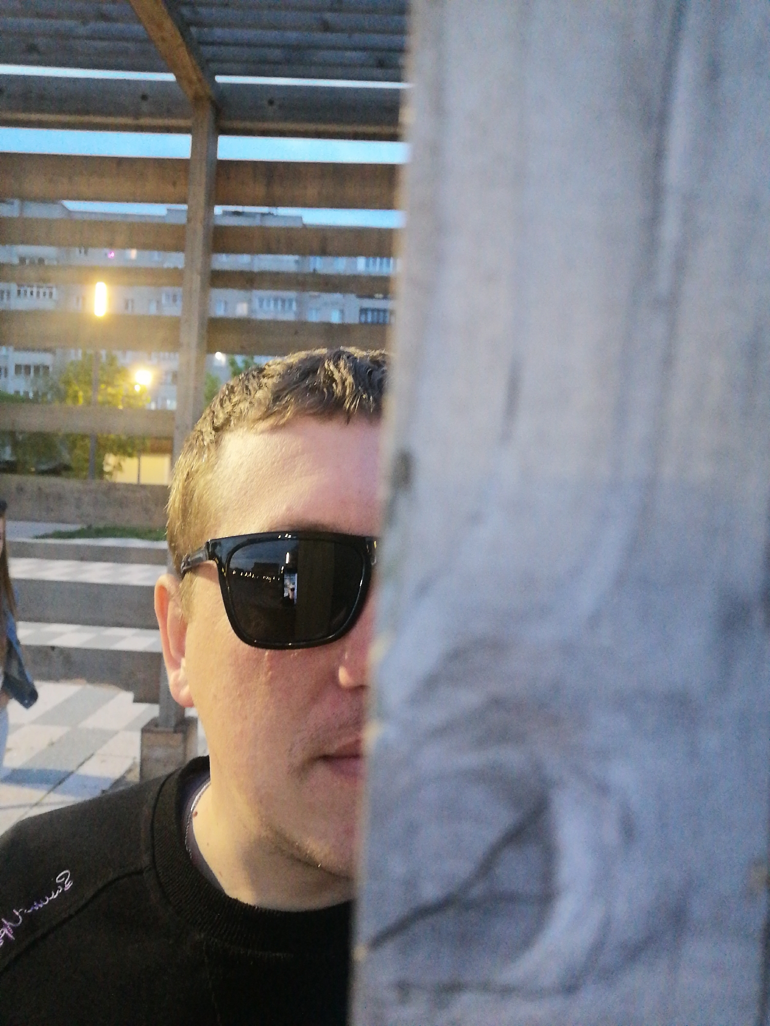 A person is partially obscured by a gray pole-like object with wood grain texture. The person is wearing black sunglasses and a black long-sleeved shirt. The background features a wooden pergola structure with horizontal slats, a building with multiple windows and visible trees, and paved steps. An individual is partially visible in the lower left of the frame. The location appears to be an urban outdoor area in daylight, but the exact city is Unknown, Unknown.