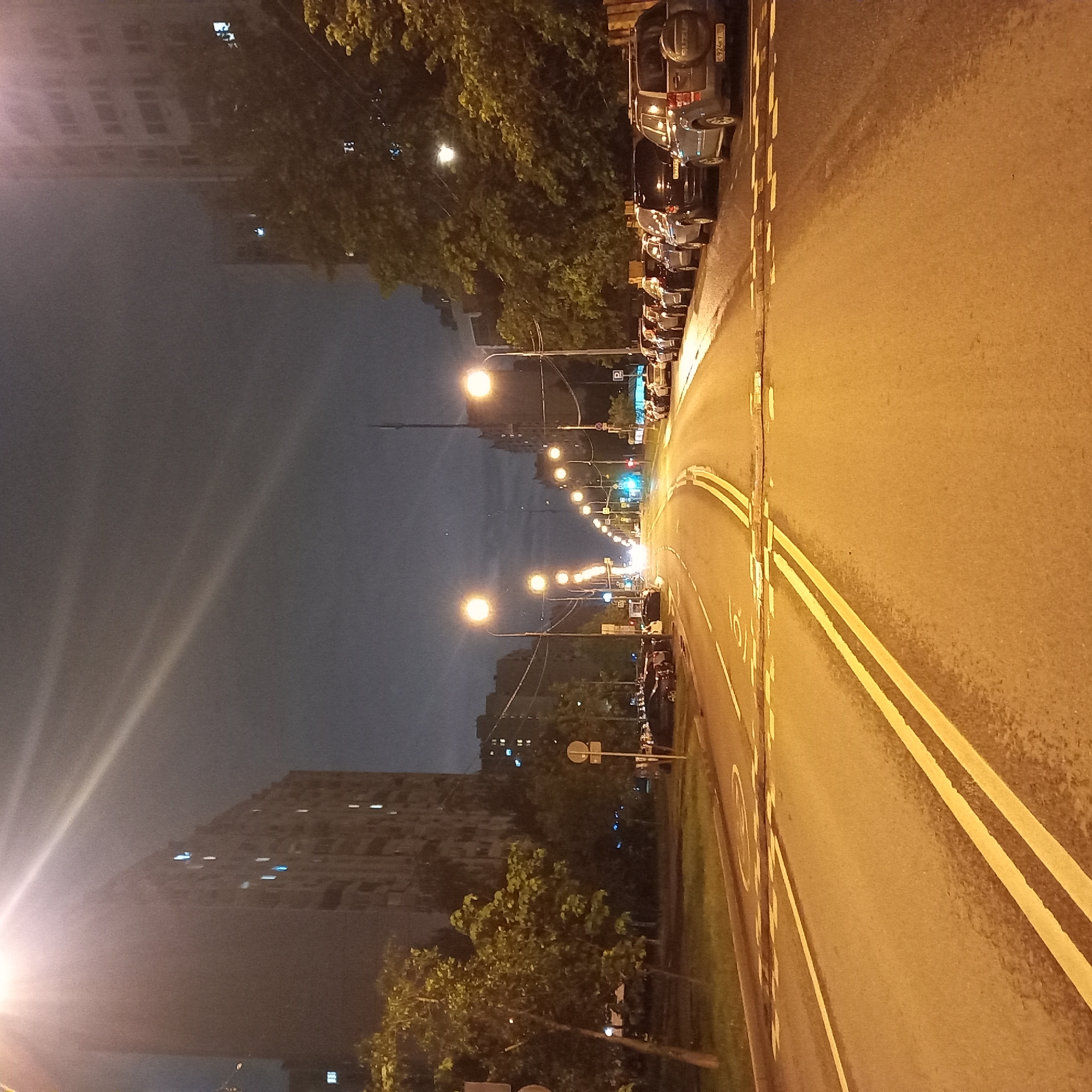 The image shows a street in Moscow, Russia, at night. The street is illuminated by streetlights, and there are cars parked along the side. The road is marked with double yellow lines and there is a building in the distance. The environment is urban.