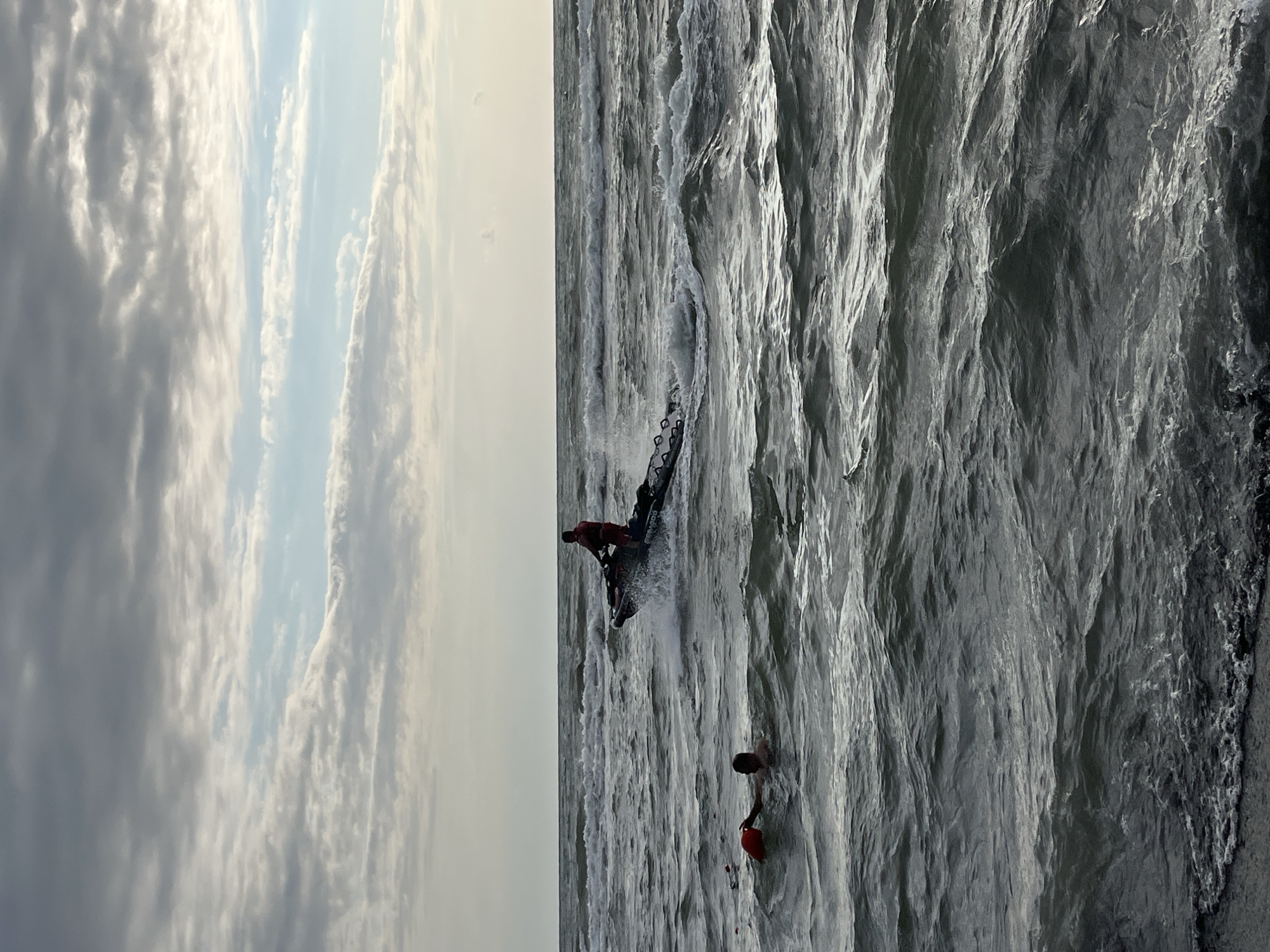 In the city of Ureki, Georgia, a lifeguard on a jet ski is approaching a person in the water. The person in the water is holding onto an orange buoy and is looking towards the approaching jet ski. The water is rough with visible waves and splashes. The sky is overcast with clouds.