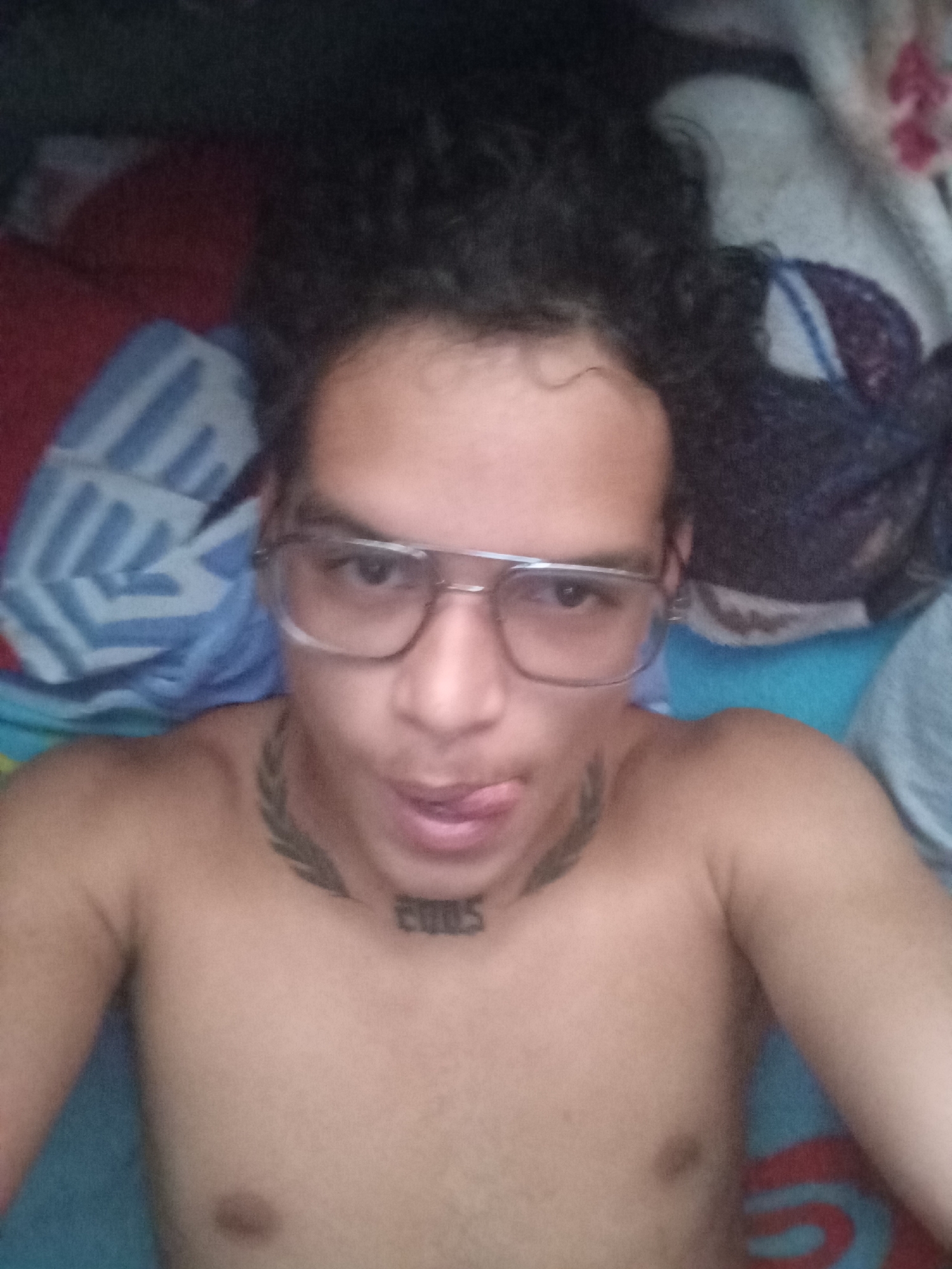 A person with curly dark hair and eyeglasses is lying shirtless on a bed. A tattoo of leaves encircles the neck, and "2005" is tattooed on the upper chest. The person is sticking their tongue out. Pillows with blue and red patterns are visible on the bed. The location and city information requested is unavailable in this image.