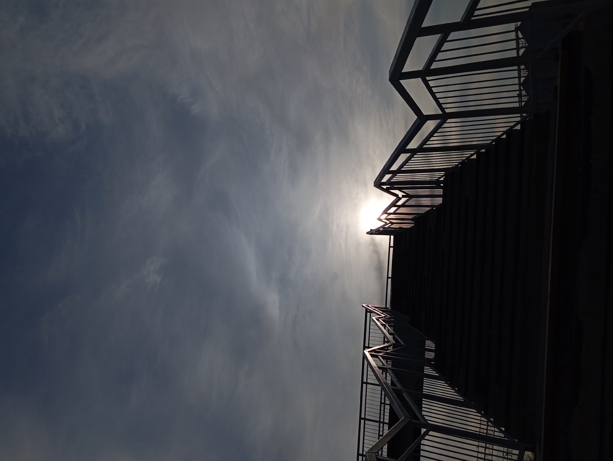 A low-angle photograph captures a metal staircase against a cloudy sky, likely taken outdoors during daylight. The metal of the staircase appears dark, nearly silhouetted, with sunlight peaking through at the top of the stairs. The sky is overcast with varying tones of gray and white clouds. The location, Yurgamysh, Russia, is not apparent from visual elements in the image. There are no people visible in the photograph.