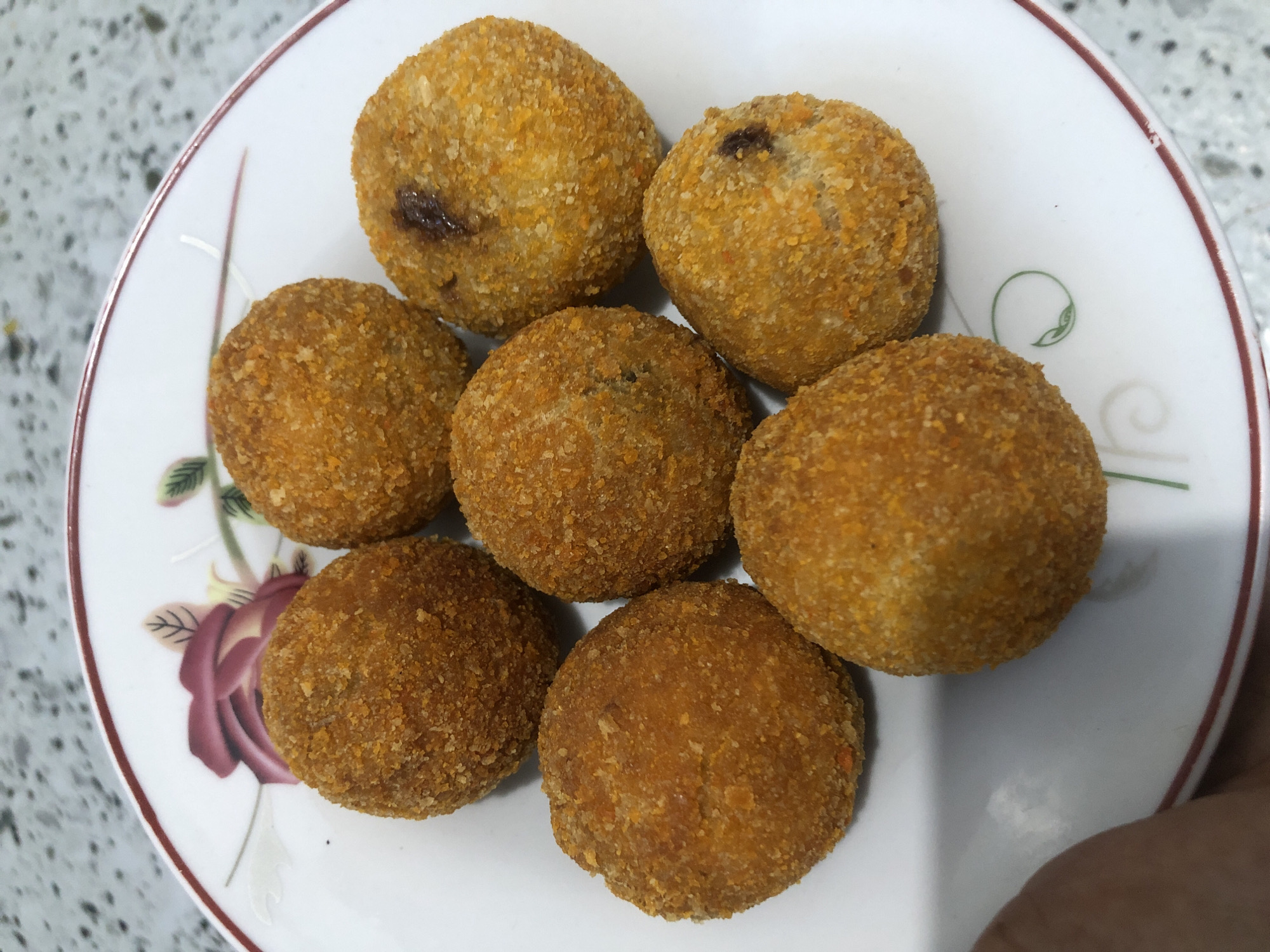 Seven golden-brown, breaded, spherical food items are arranged on a white ceramic plate with a floral design and a red rim. The food items appear to be deep-fried and have a crispy texture. Some of the items have dark spots visible on their surface, suggesting a filling. The plate rests on a speckled, light-colored surface. The image's metadata indicates the location as Gampongasanlhoksukon Barat, Indonesia. A hand is partially visible in the lower left corner, holding the edge of the plate.