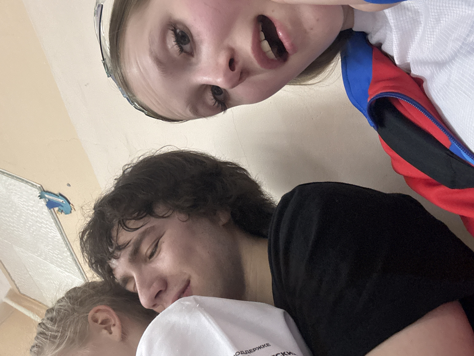 The image shows three people indoors. A young woman with blonde hair and wearing a white and blue/red tracksuit is in the upper part of the frame, looking into the camera with an open mouth. Below her is a young man with dark curly hair, resting his head on the shoulder of another person, who is only partially visible at the bottom left. The background is a plain wall.
Given the tracksuit's colours (white, blue, and red), it is possible this image was taken in Russia. Without other evidence, I cannot verify its presence in the city of Usolye.