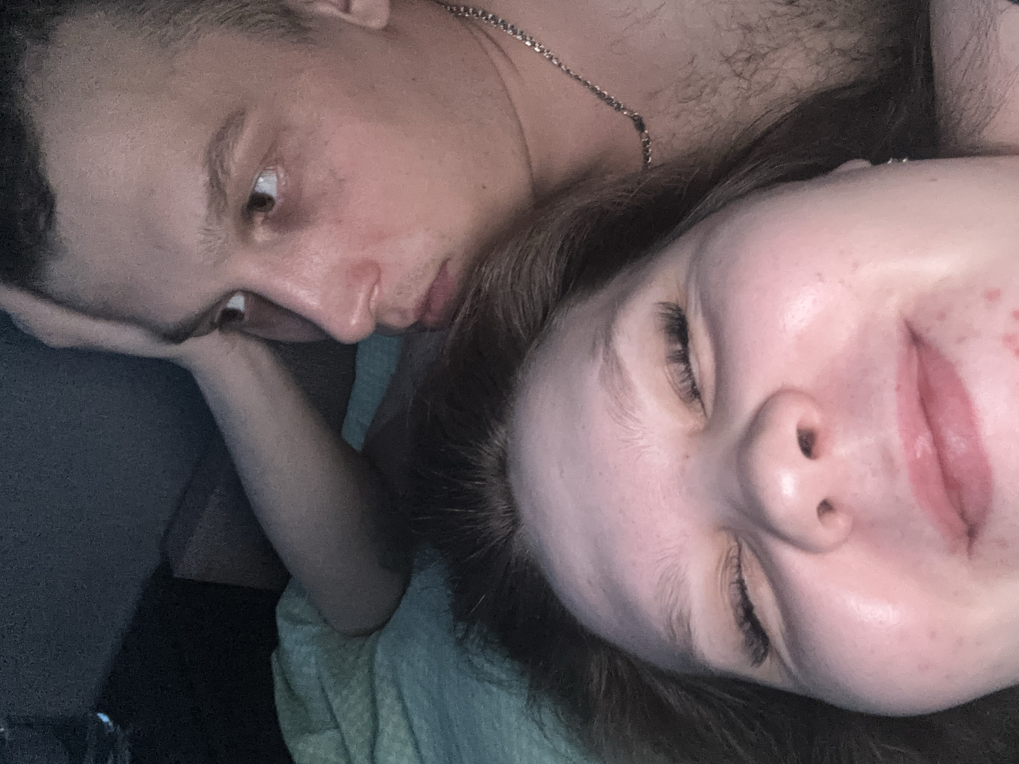 A dimly lit, close-up photograph shows two people lying down. A fair-skinned male with short dark hair is partially visible, his head resting on his hand. His eyes are open, looking to the side. A silver chain is visible around his neck. Next to him, a fair-skinned female with dark hair lies with her eyes closed. The environment appears to be an indoor space, likely a bedroom or living room. There is a blanket underneath the individuals and the location cannot be verified to be in Chaikovsky, Russia.