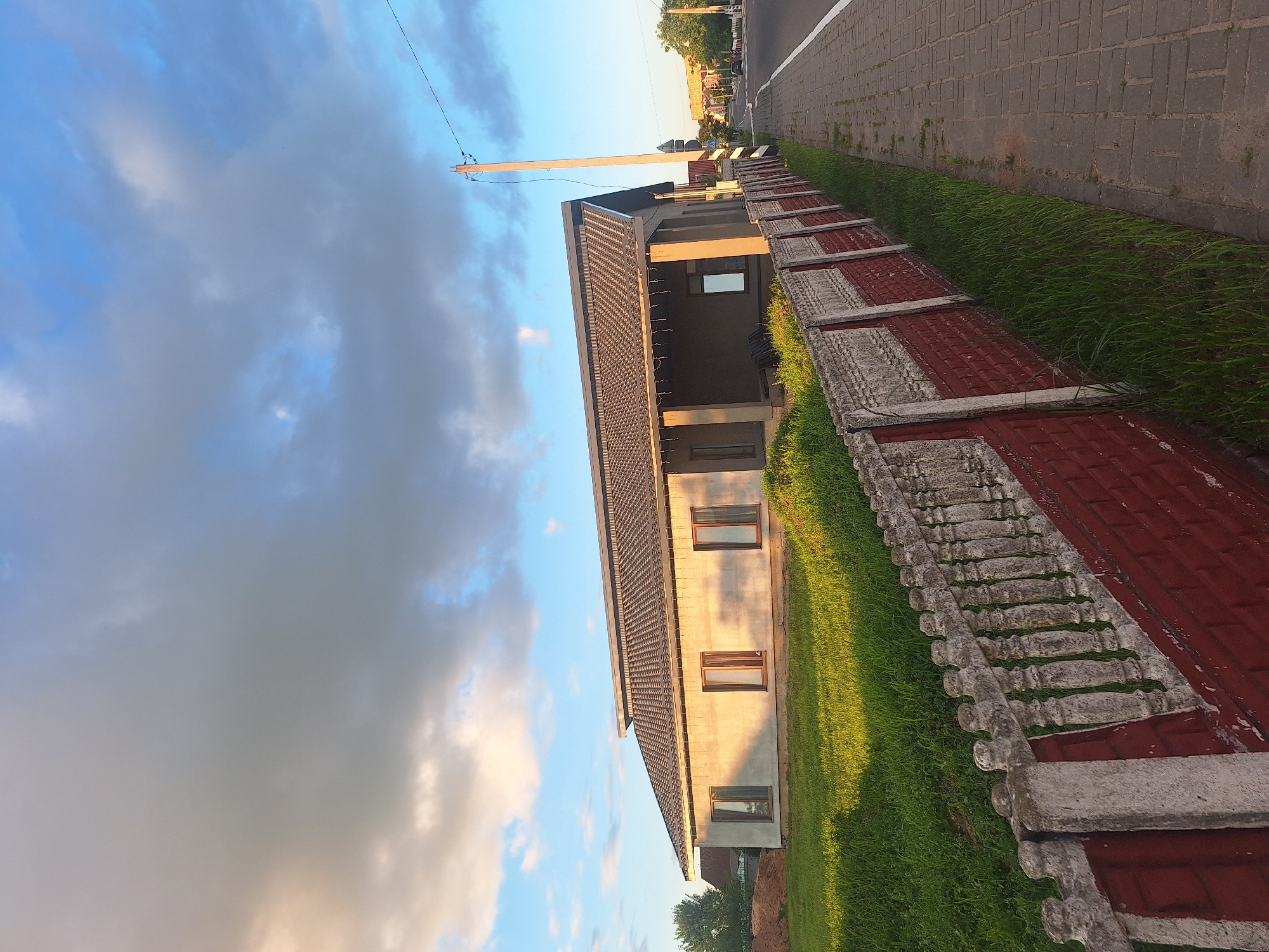 The image captures a street-side view of a single-story house under construction, located in the Kapylskiy Selski Soviet, Belarus. The house is a concrete structure with a dark tiled roof and several rectangular windows. In front of the house, a lawn is partially illuminated by warm, low-angle sunlight, casting long shadows. A decorative fence, made of red brick and concrete balusters, runs along the property line adjacent to a paved sidewalk. A single wooden utility pole stands on the right side of the frame, with wires extending across the sky. The sky is a mix of blue and gray clouds, with the sun's light suggesting it is either early morning or late afternoon. In the distance, other buildings and trees are visible, along with a street where a car is present. The overall scene depicts a semi-urban or rural residential area during a period of ongoing construction.