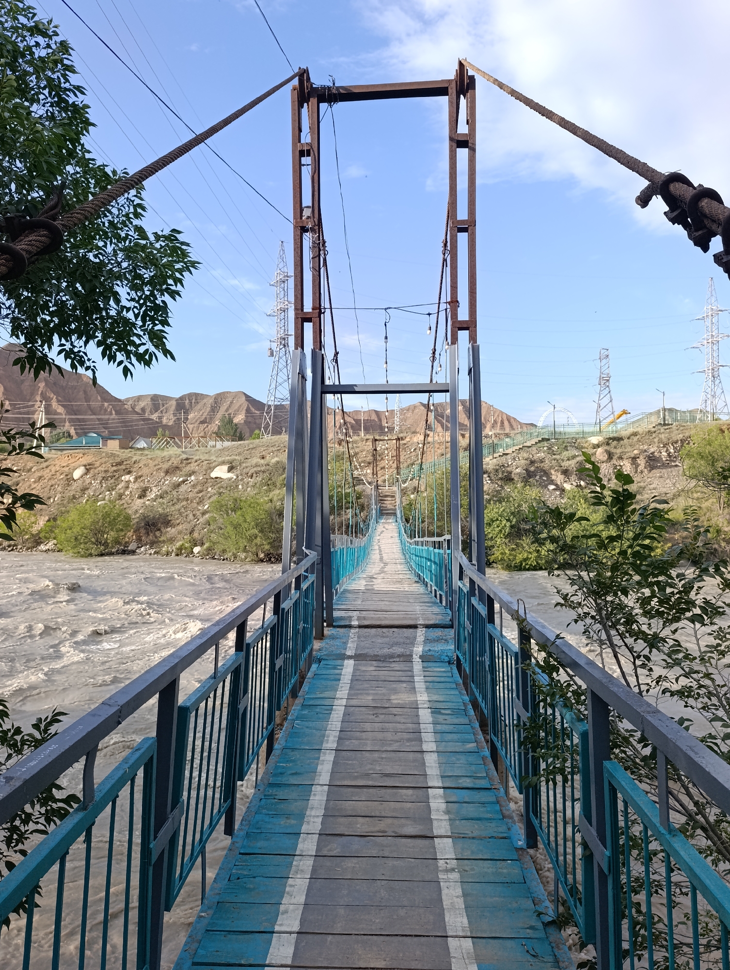 An eye-level, vertical, medium shot captures a weathered pedestrian suspension bridge, painted blue and white, spanning a muddy, turbulent river in Naryn City, Kyrgyzstan. The bridge's wooden planks are worn, and its metal railings, painted blue, show signs of rust. On either side of the bridge, dense green foliage borders the walkway. The river, a churning mass of light brown water, rushes beneath the structure. In the background, steep, barren hills rise under a clear, pale blue sky dotted with wispy white clouds. Power lines and tall transmission towers are visible against the landscape, suggesting the presence of infrastructure near the natural terrain. The scene is devoid of people, with the focus entirely on the bridge and its surroundings.