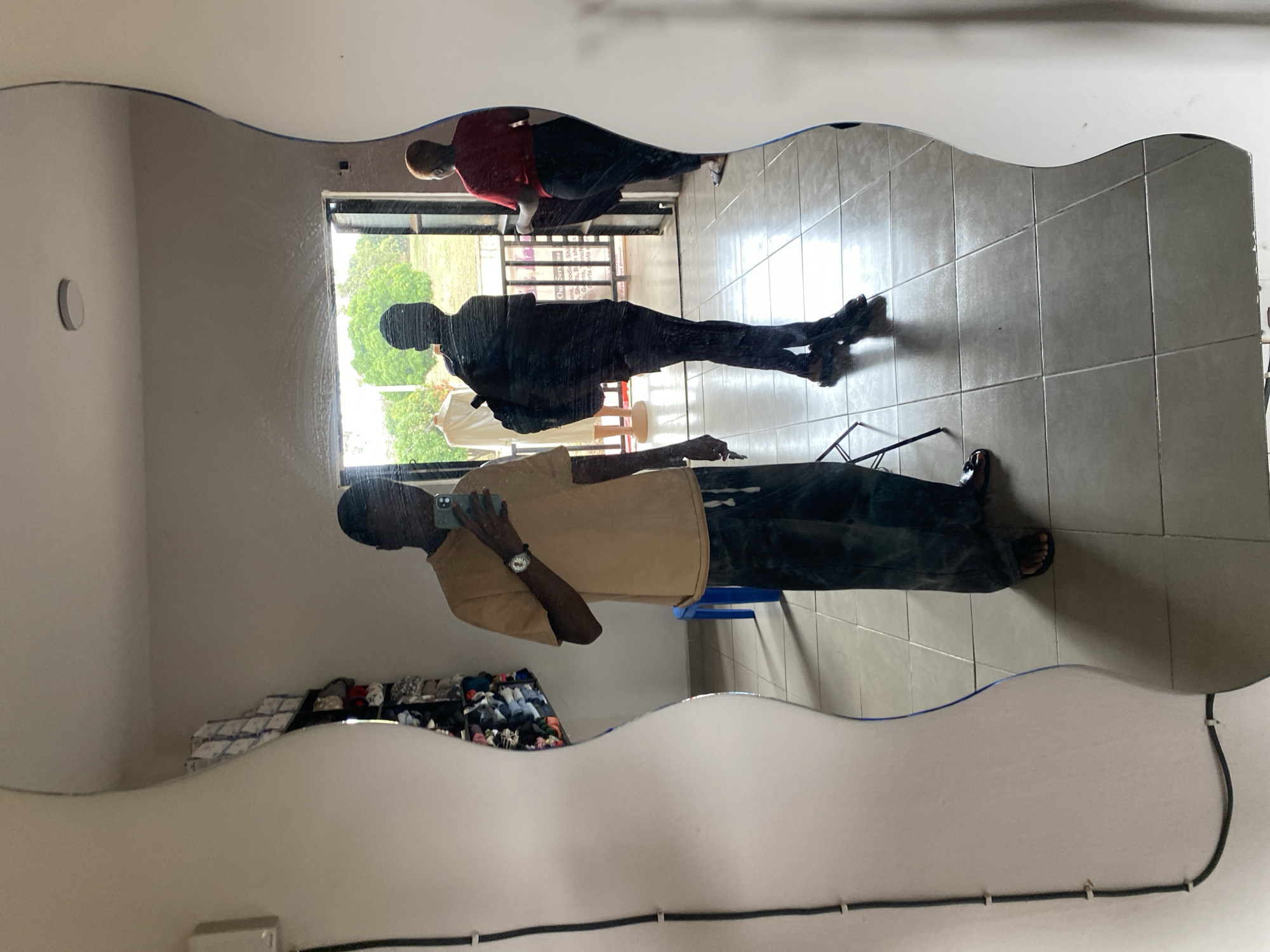 The media file displays a mirror reflection of an indoor scene, oriented vertically and appearing rotated 90 degrees clockwise. The primary subject is a person taking a selfie, reflected in the foreground, wearing a beige short-sleeved top and dark pants, holding a smartphone with a light green case. A watch is visible on their left wrist.
Behind the person taking the photo, two other figures are reflected in the mirror. One, closer to the foreground, appears as a dark silhouette wearing a dark top and pants. The other, further back and closer to a large window, is seen wearing a red top and dark pants.
The mirror itself has a distinctive wavy, irregular edge along its top. The reflection reveals a tiled floor in a light color. Through the window reflection, bright daylight illuminates green trees and foliage, indicating an outdoor view during the day. Inside the reflected room, a storage unit or rack containing numerous shoes or folded clothing items is visible in the background. A black cable is secured to the wall with white ties along the bottom edge of the mirror's reflection. A round, white object is mounted on the wall in the reflection. Faint, partially obscured text, possibly "ssage" or "Shop" from a sign, can be seen in the window's reflection. The setting is in Bukuru, Nigeria.