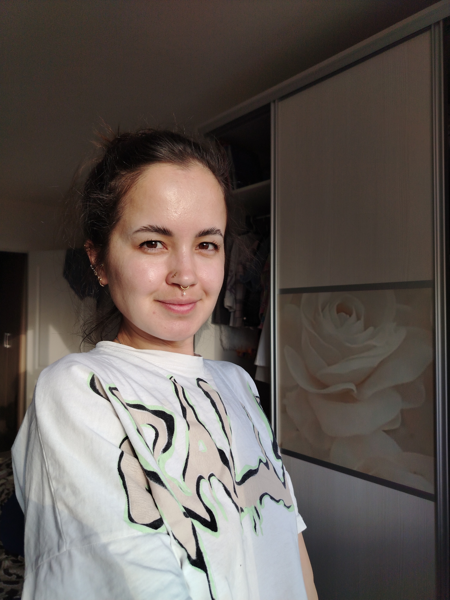 A young adult female is posing for a selfie indoors. She has brown hair pulled back in a messy bun, brown eyes, and fair skin. She is wearing a white T-shirt with an abstract design of black and beige shapes outlined in light green. She has multiple piercings on her left ear and a ring in her nose.
Behind her is a wardrobe with sliding doors, one of which has a large image of a white rose. The background is slightly out of focus, but it appears to be a room in a residential setting. The scene is lit by natural light, creating shadows on the wall to the left of the subject.
The city is not visible.
